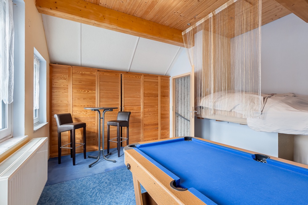Zimmer mit Billardtisch, erhöhtem Bett, kleinem Tisch und zwei Stühlen, sichtbarem Holzbalken und Fenstern.