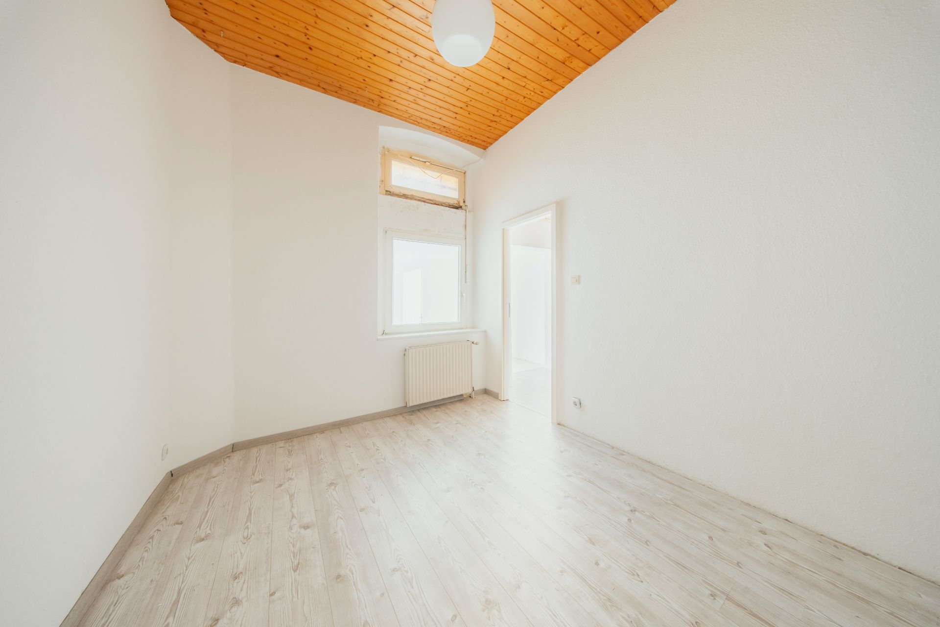 Helles Zimmer mit Holzdecke, hellem Boden, Fenster, Heizung und Tür zu einem weiteren Raum.