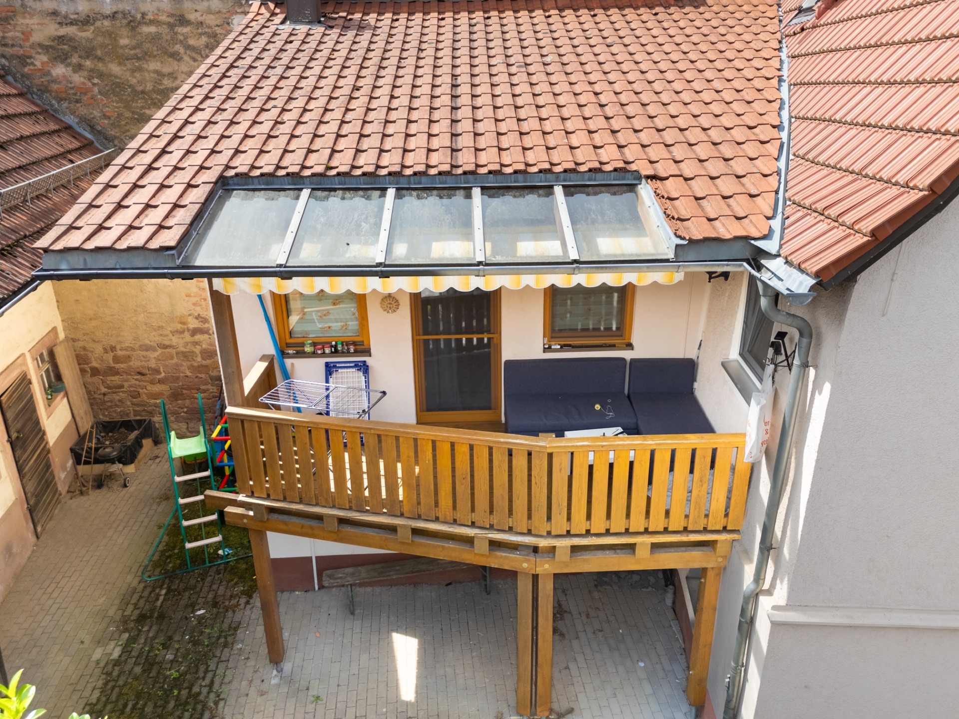 Überdachter Balkon mit Holzgitter, Sofa, Wäscheständer und Zugang durch zwei Fenster.