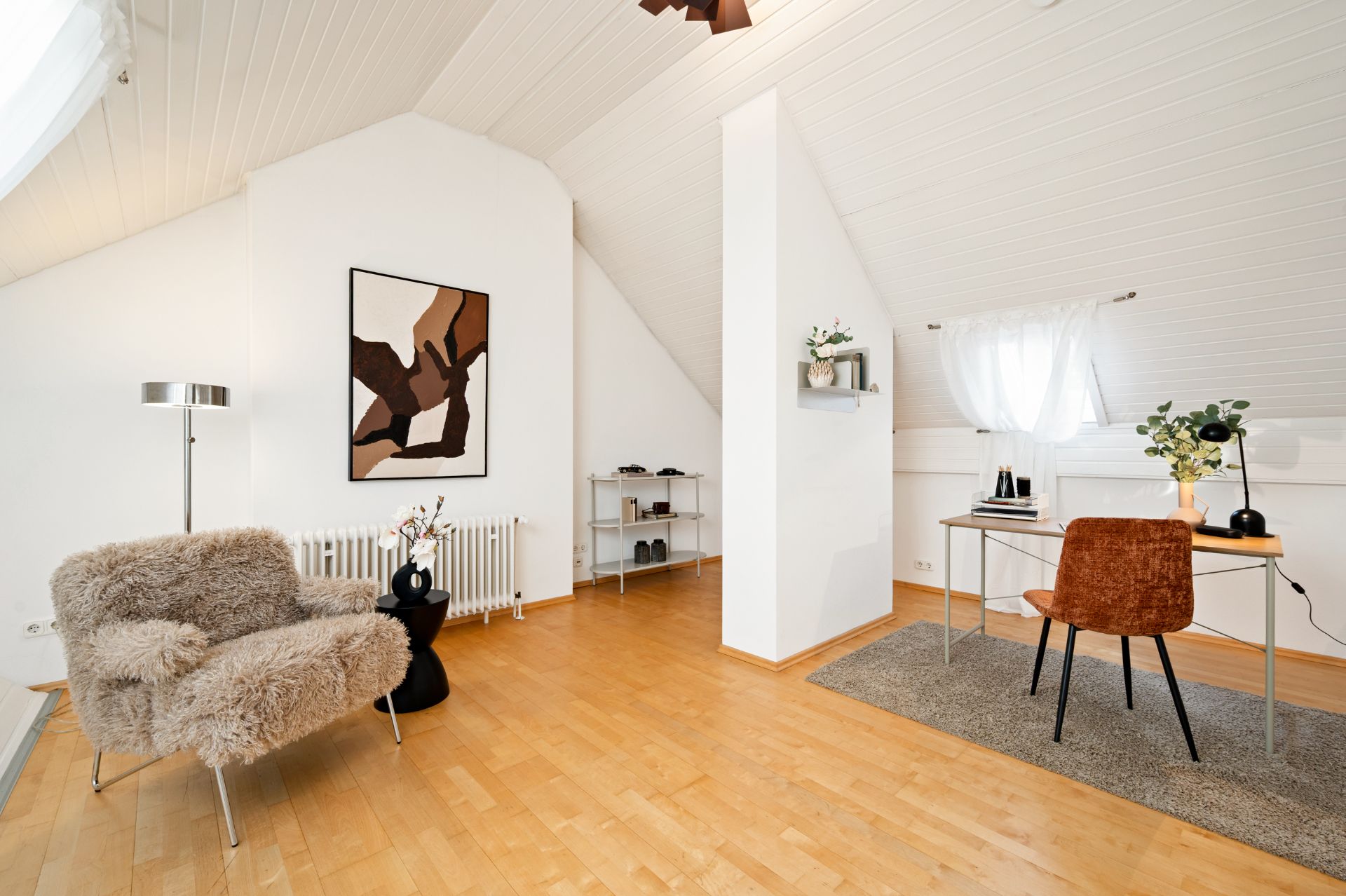 Helles Dachzimmer mit Schreibtisch, Sessel, Wandbild und Regal auf Holzfußboden.