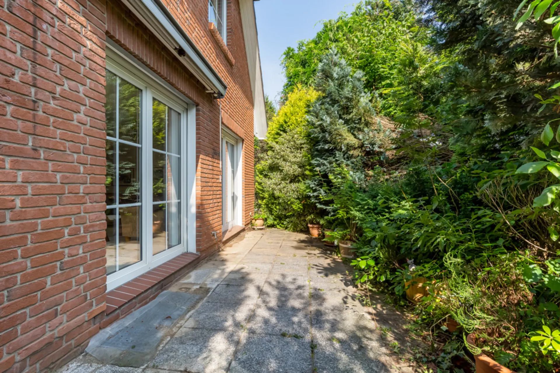 Terrasse mit Steinfliesen neben einem roten Ziegelhaus, umgeben von vielen grünen Pflanzen.