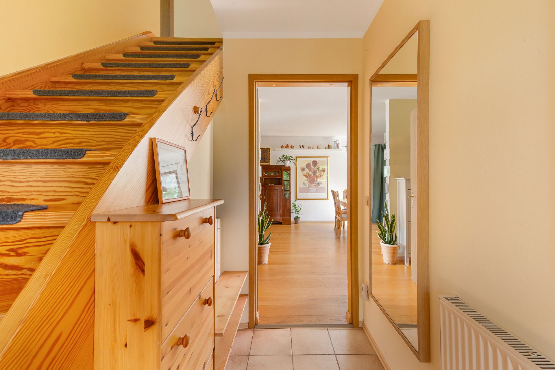 Hausflur mit Holztreppe, Kommode, großem Spiegel und Blick ins Wohnzimmer mit Pflanzen.