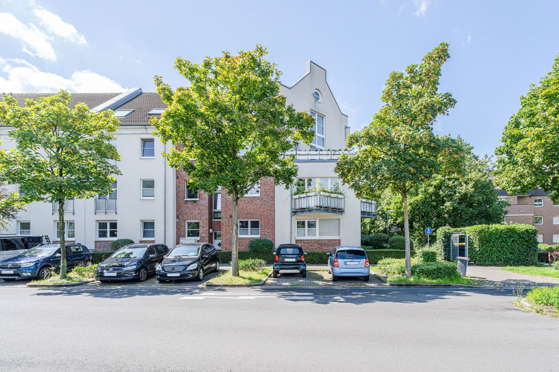 Mehrfamilienhaus mit Balkonen, Bäumen davor und Autos auf Parkplätzen an der Straße.