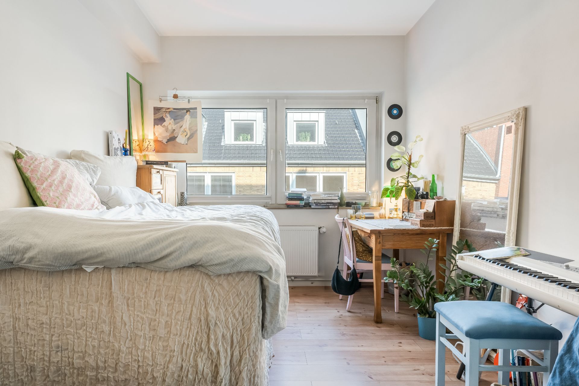 Helles Schlafzimmer mit Bett, Schreibtisch, Spiegel, Pflanzen und einem Keyboard am Fenster.