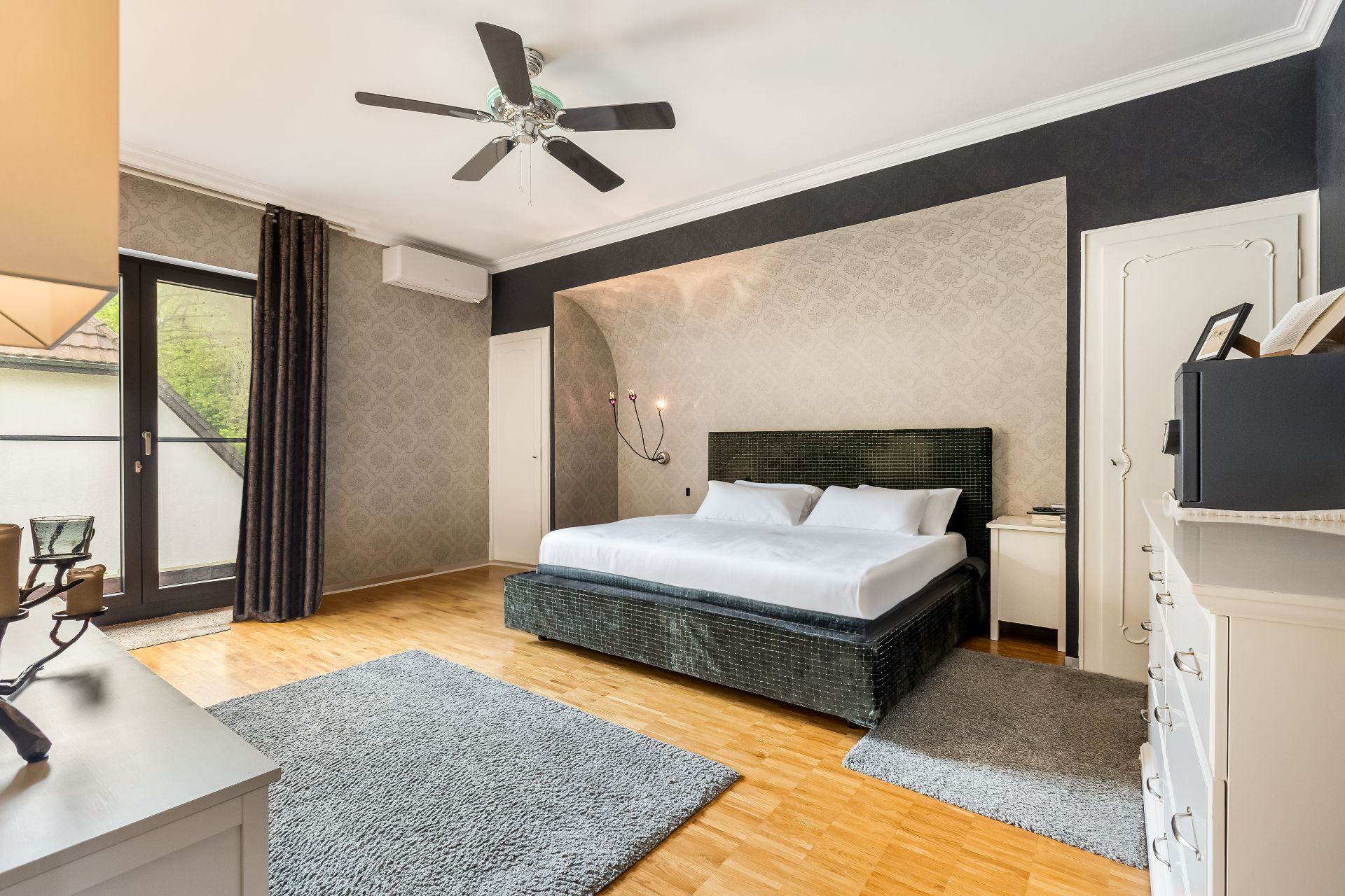 Modernes Schlafzimmer mit großem Bett, Deckenventilator, Teppichen und Zugang zum Balkon.