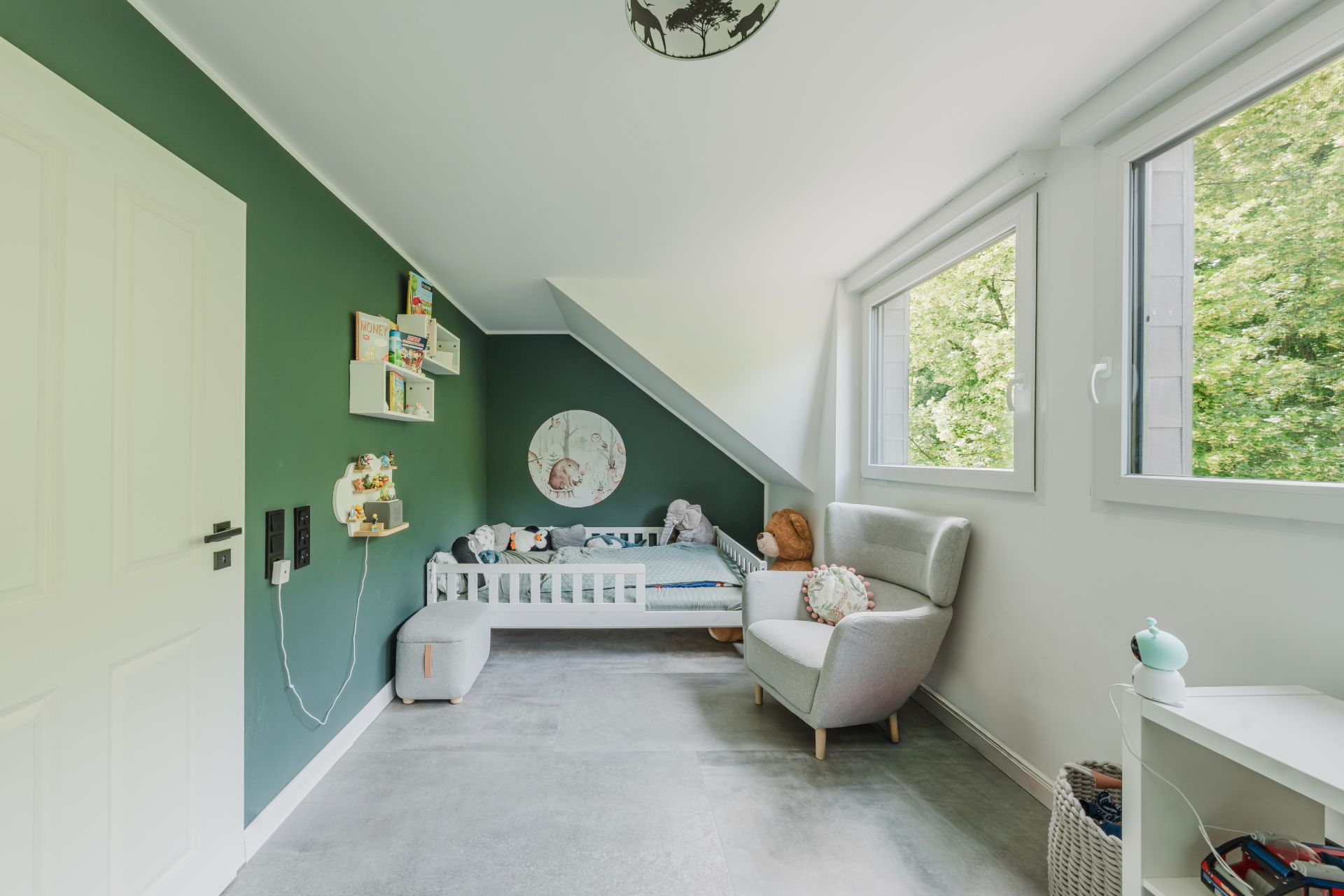 Helles Kinderzimmer mit grün gestrichener Wand, Bett, Sessel und großen Fenstern.