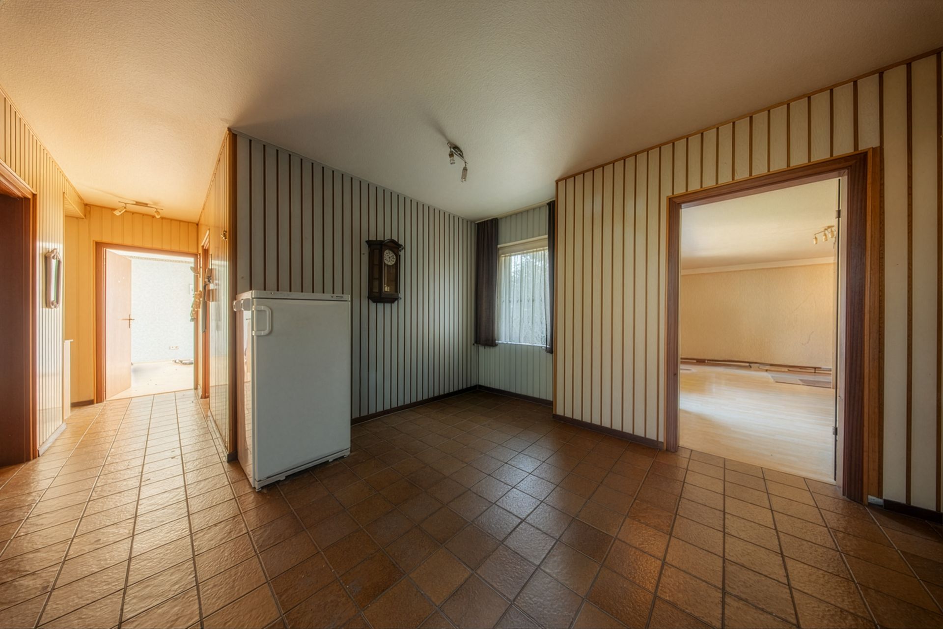 Flur mit braunen Fliesen, Kühlschrank, Wanduhr und Zugang zu weiteren Zimmern.