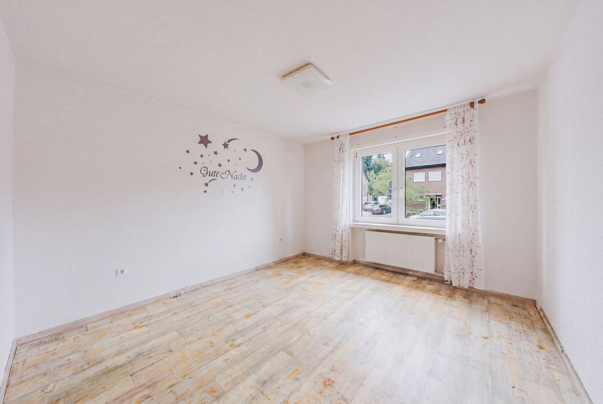 Leerer, heller Raum mit Holzfußboden, Wandtattoo und Fenster mit weißen Vorhängen.