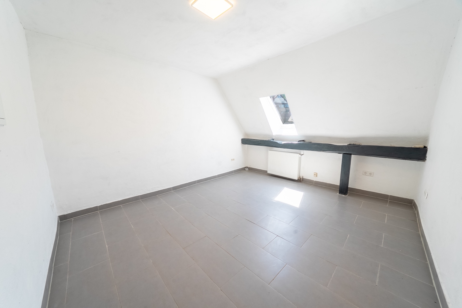 Leerer Raum mit schräger Decke, grauem Fliesenboden und kleinem Fenster.