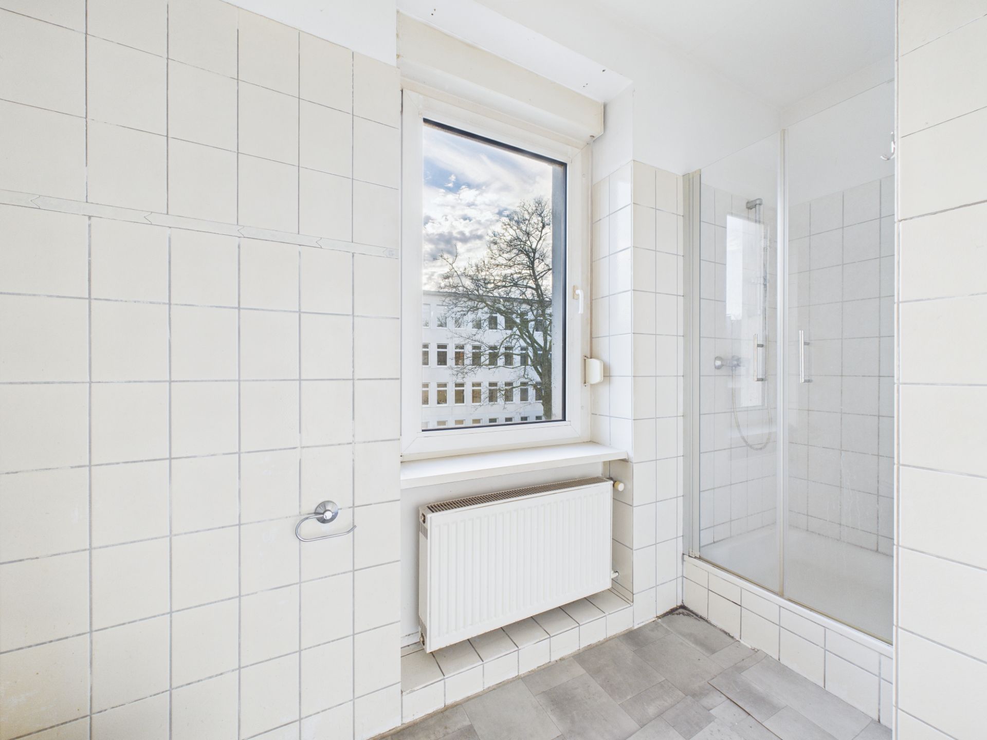 Helles Badezimmer mit weiß gefliesten Wänden, Fenster, Heizung und Glasdusche.