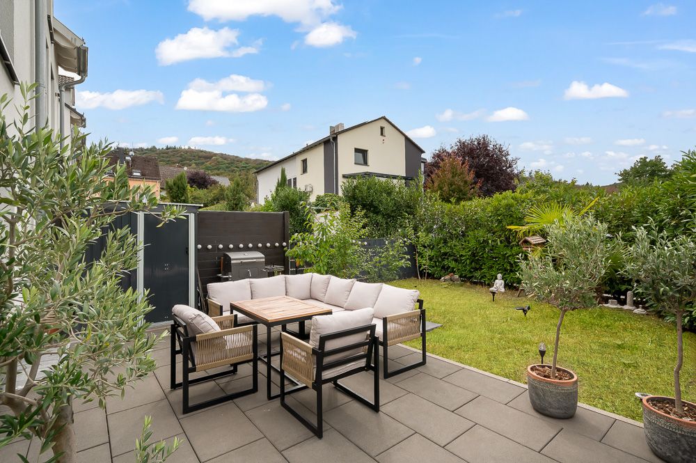 Terrasse mit Sitzgruppe, Tisch und Pflanzen, angrenzend an einen Garten mit Rasen und Hecken.