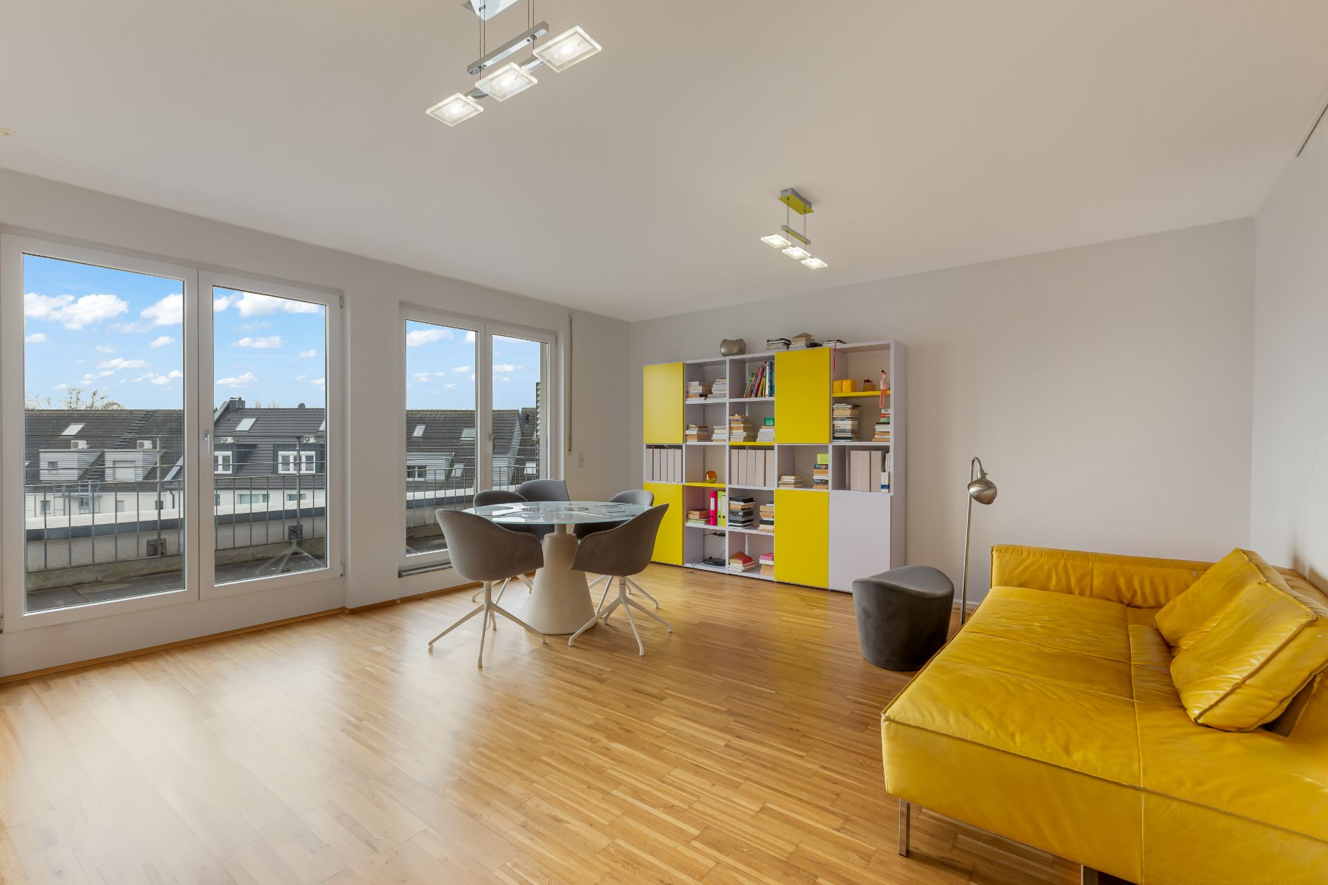 Helles Wohnzimmer mit gelbem Sofa, großem Fenster, Tisch mit Stühlen und Regal mit gelben Türen.