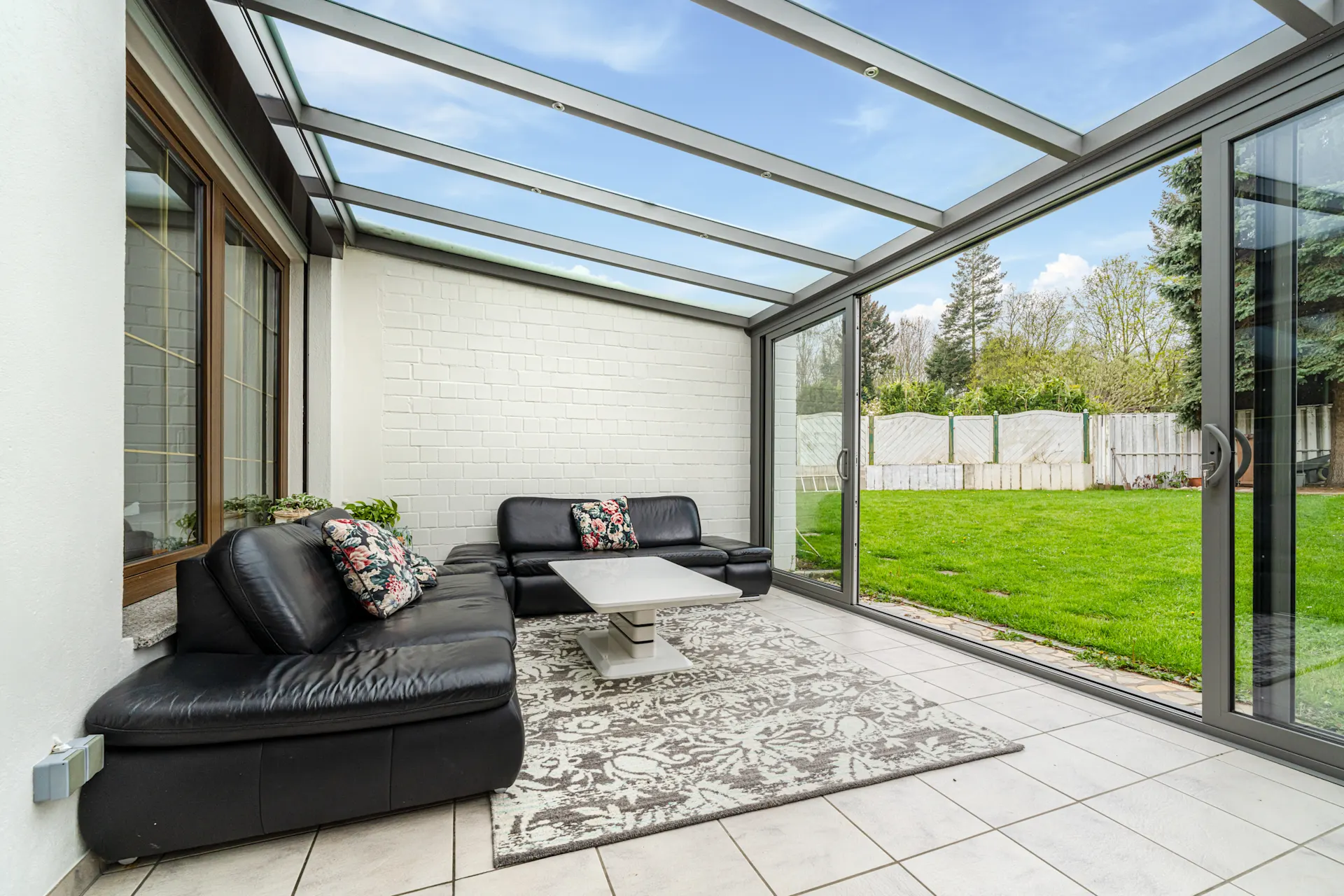 Wintergarten mit Glasdach, schwarzen Sofas, Teppich und Blick in den Garten.