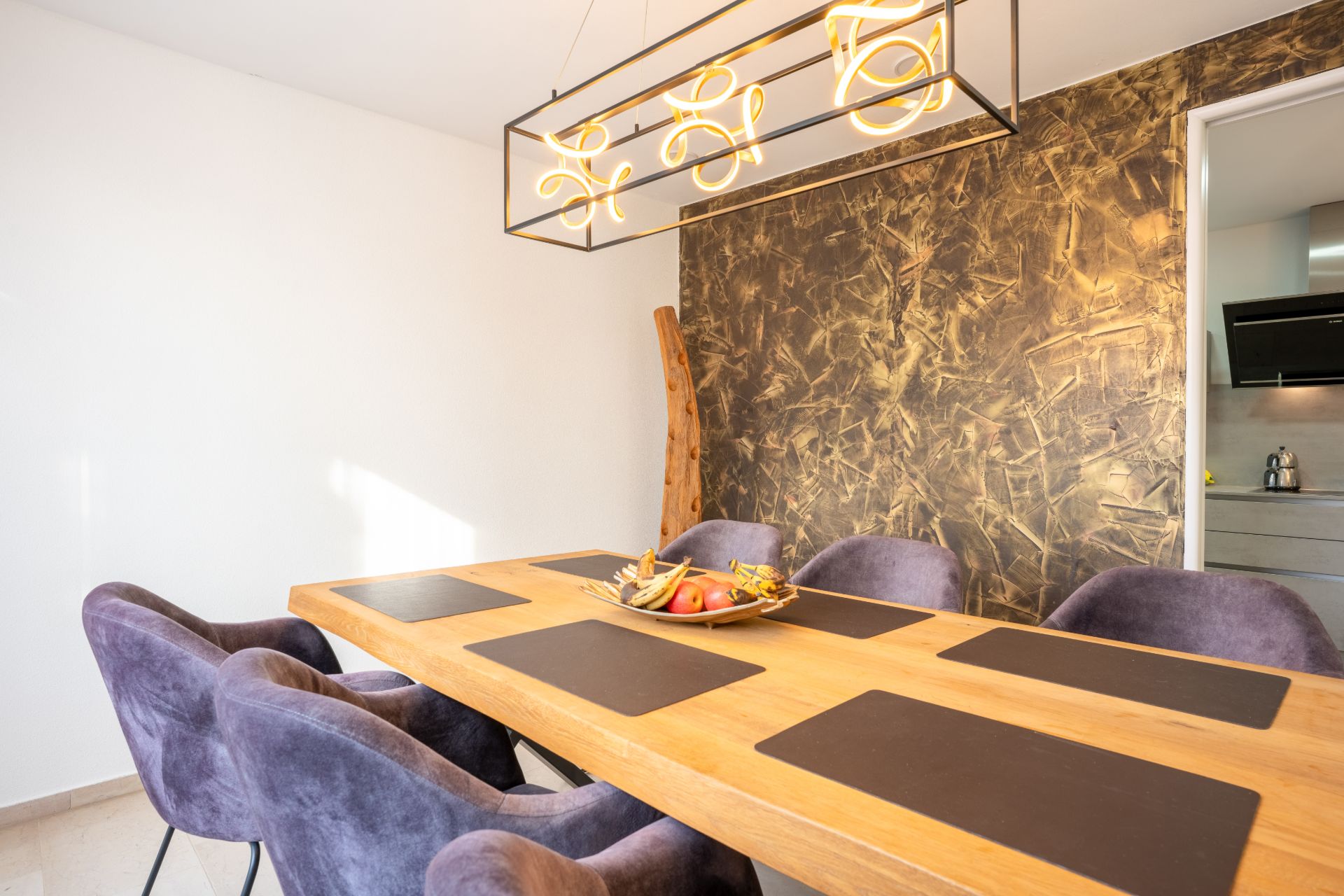 Esszimmer mit Holztisch, sechs violetten Stühlen und moderner Hängelampe an strukturierter Wand.