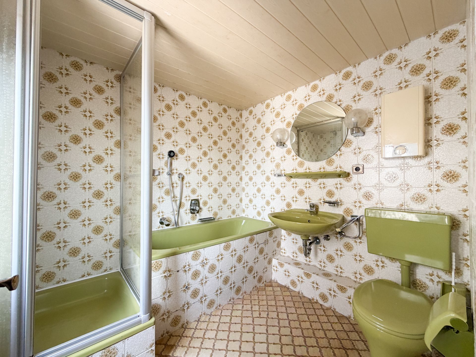 Badezimmer mit grüner Badewanne, WC und Waschbecken, gemusterten Fliesen und rundem Spiegel.