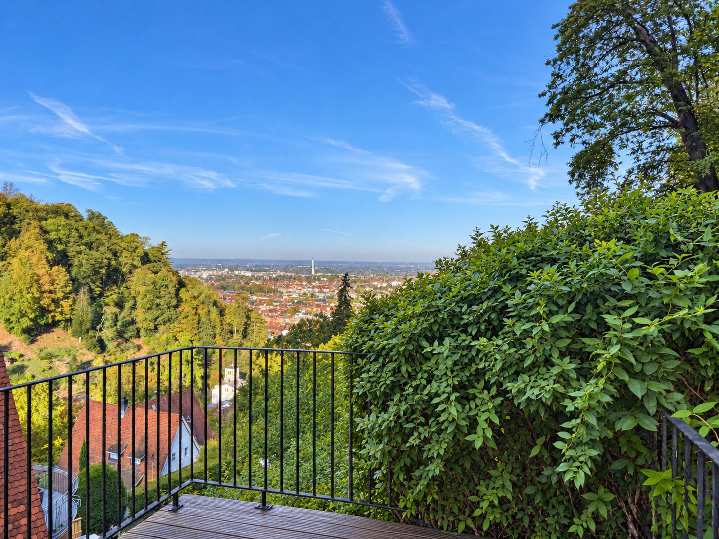 Balkon mit Metallgeländer, Blick auf grüne Hügel und Stadt im Hintergrund bei klarem Himmel.