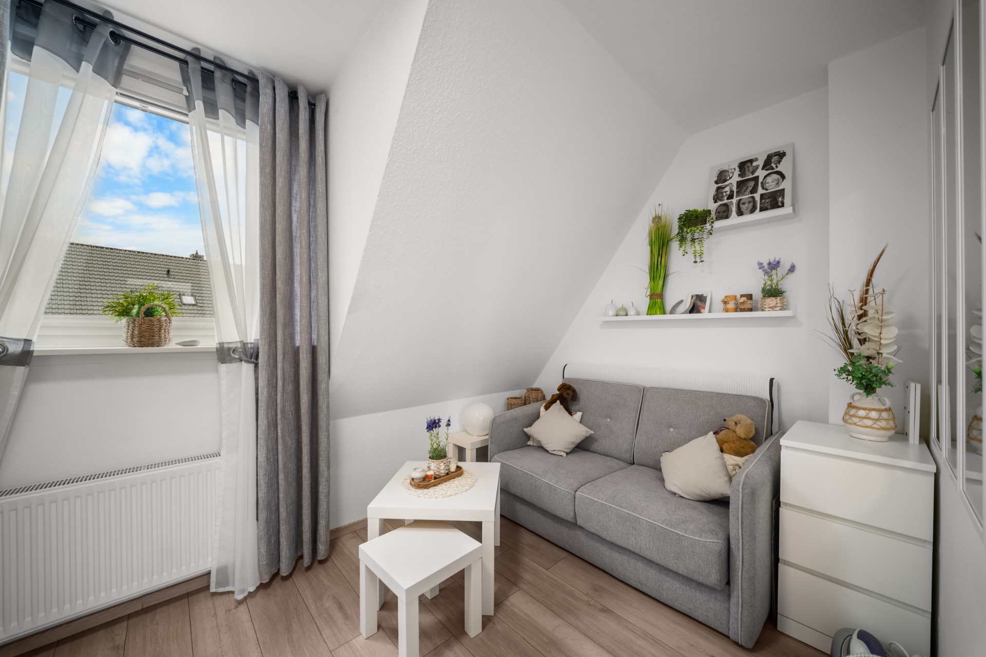 Helles Wohnzimmer mit grauem Sofa, Couchtisch, Pflanzen und großem Fenster mit Vorhängen.