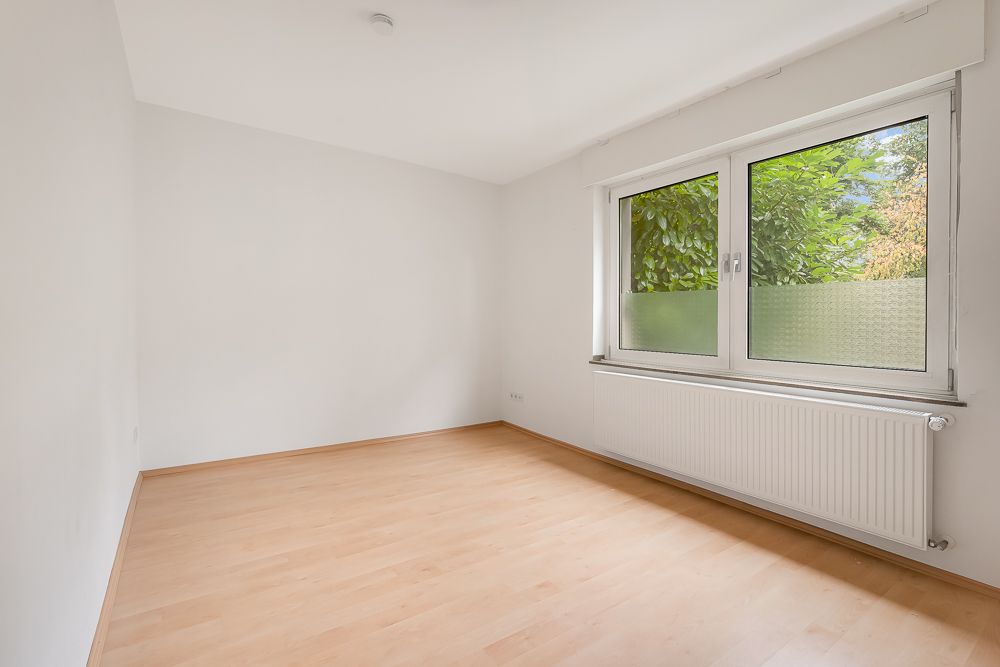 Heller, leerer Raum mit hellem Holzfußboden und großem Fenster mit Blick ins Grüne.
