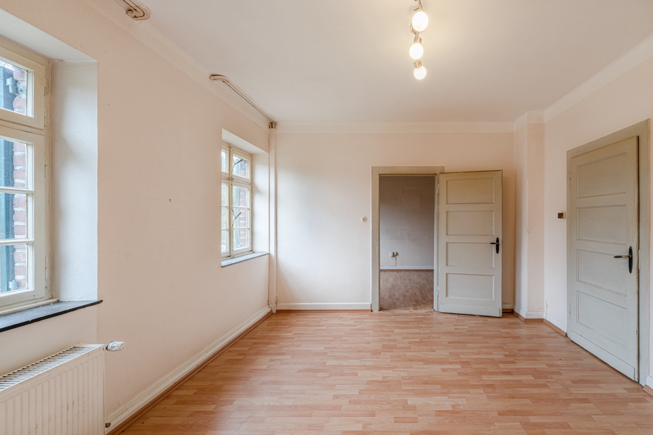 Helles, leeres Zimmer mit Holzfußboden, zwei Fenstern und zwei geöffneten Türen.