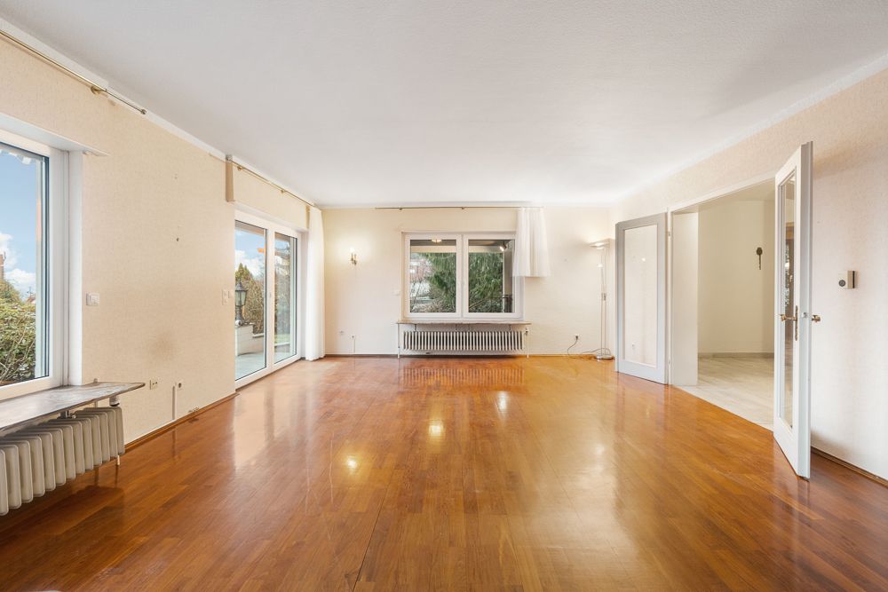Großes, leeres Wohnzimmer mit Holzfußboden und großen Fenstern, Türen führen nach draußen und in einen Nebenraum.