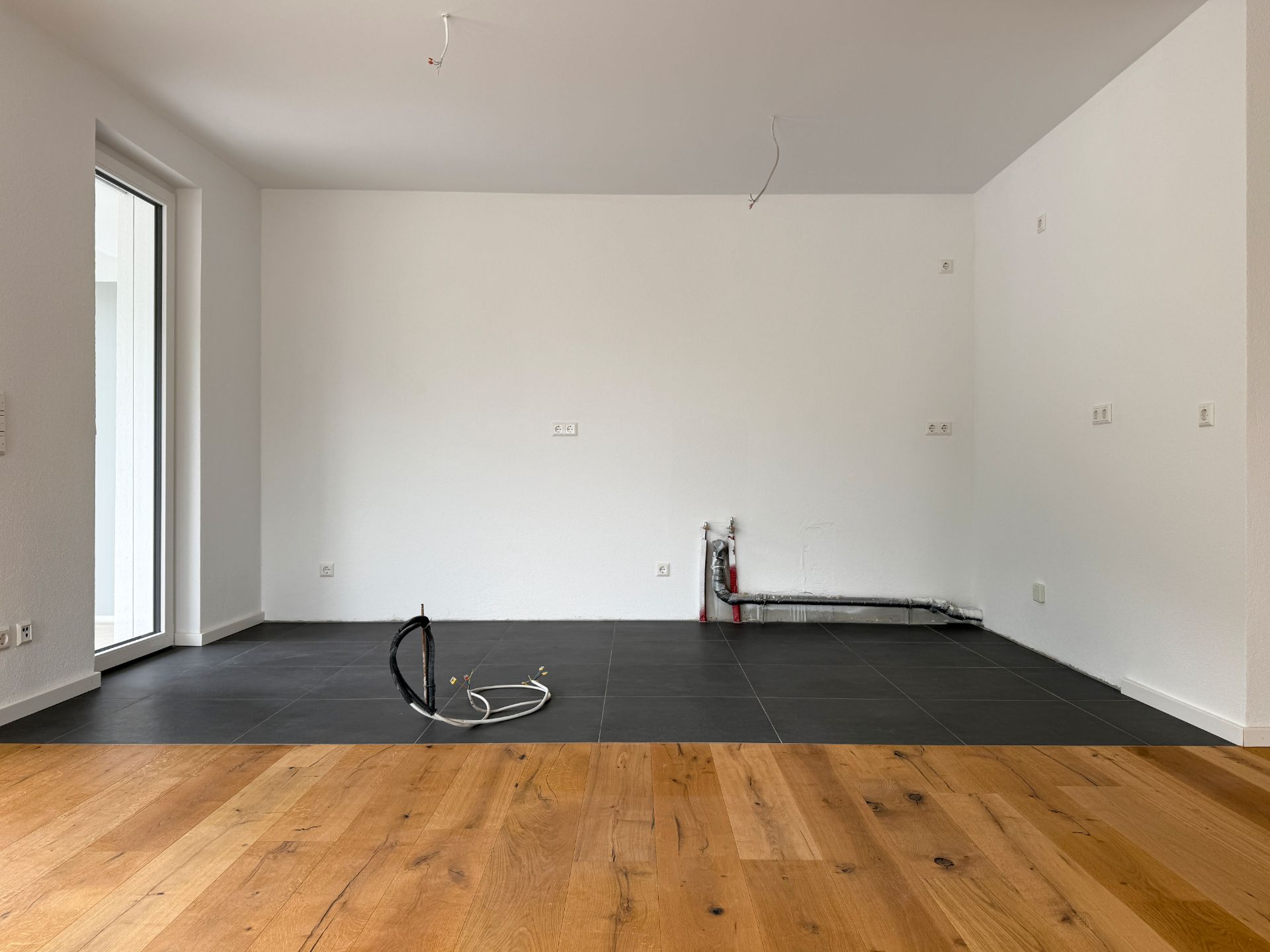 Leerer Raum mit hellem Holzboden und schwarzem Fliesenbereich, Kabel am Boden, weiße Wände.