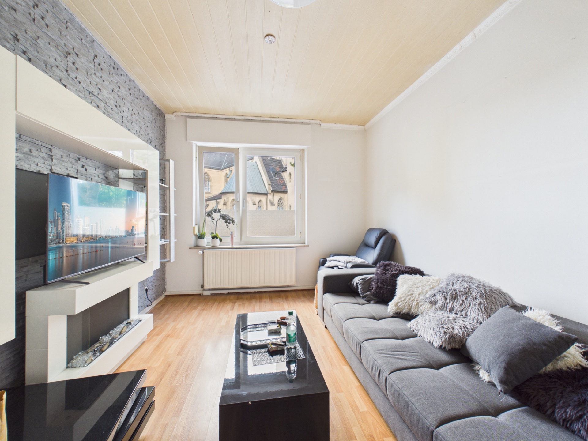 Wohnzimmer mit grauem Sofa, Couchtisch, Fernseher an Steinwand und großem Fenster.