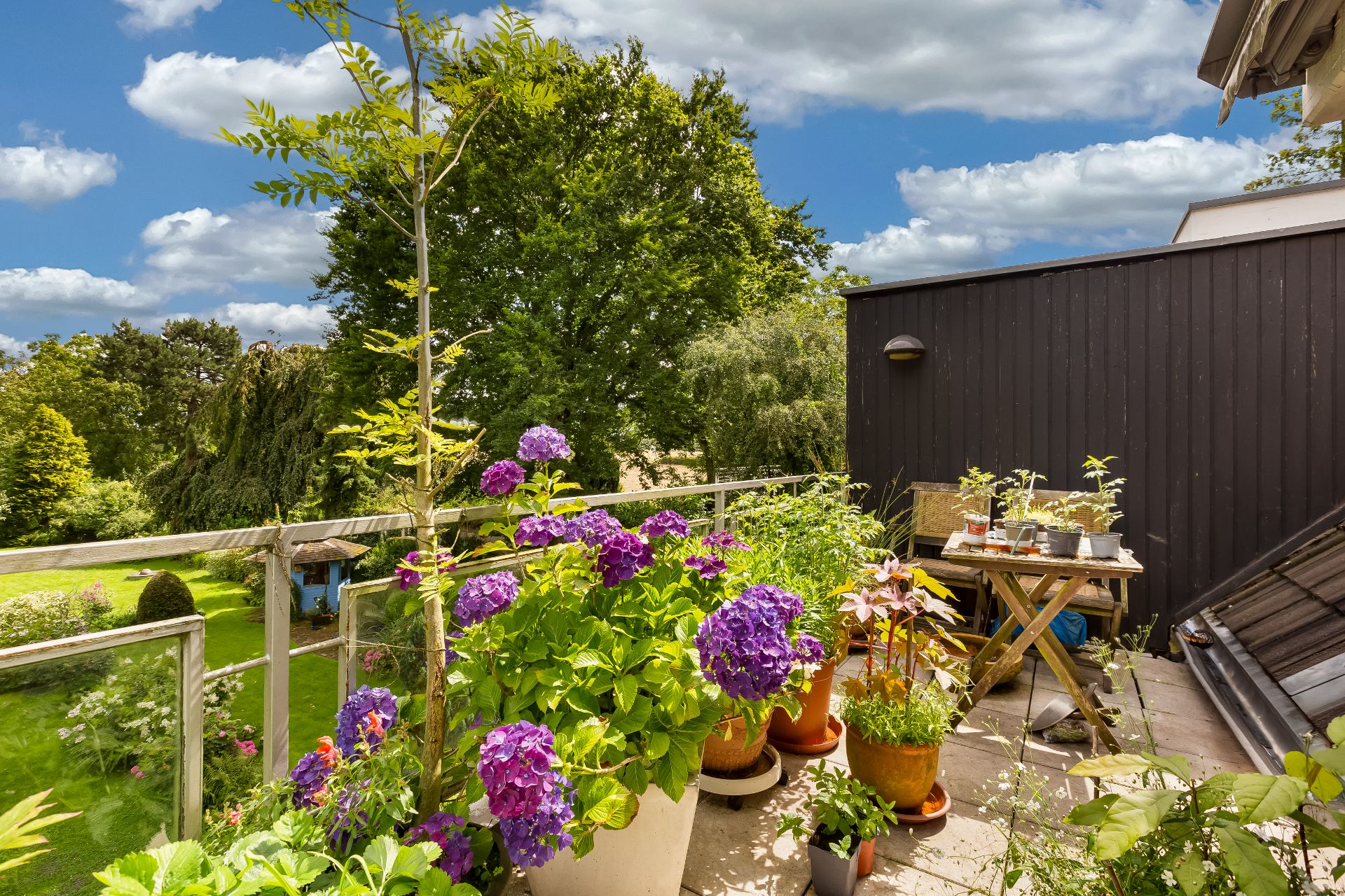 Balkon mit vielen Blumentöpfen und Blick auf einen grünen Garten mit Bäumen.
