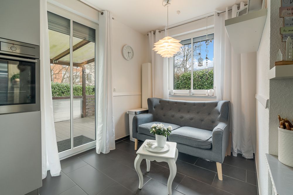 Kleines Wohnzimmer mit grauem Sofa, weißem Tisch und Fenster, daneben Glastür zur Terrasse.