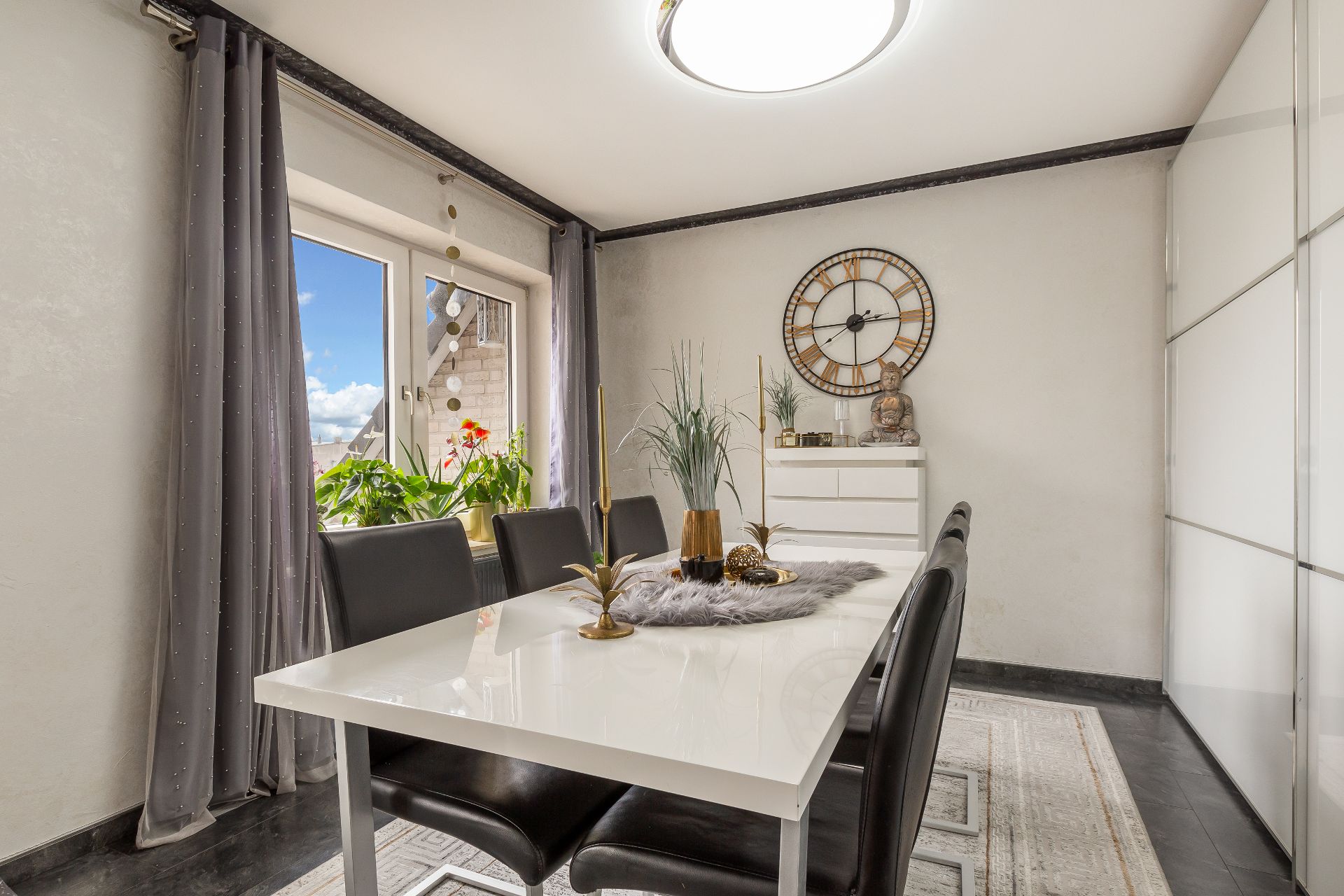 Esszimmer mit weißem Tisch, schwarzen Stühlen, Pflanzen am Fenster und großer Wanduhr.