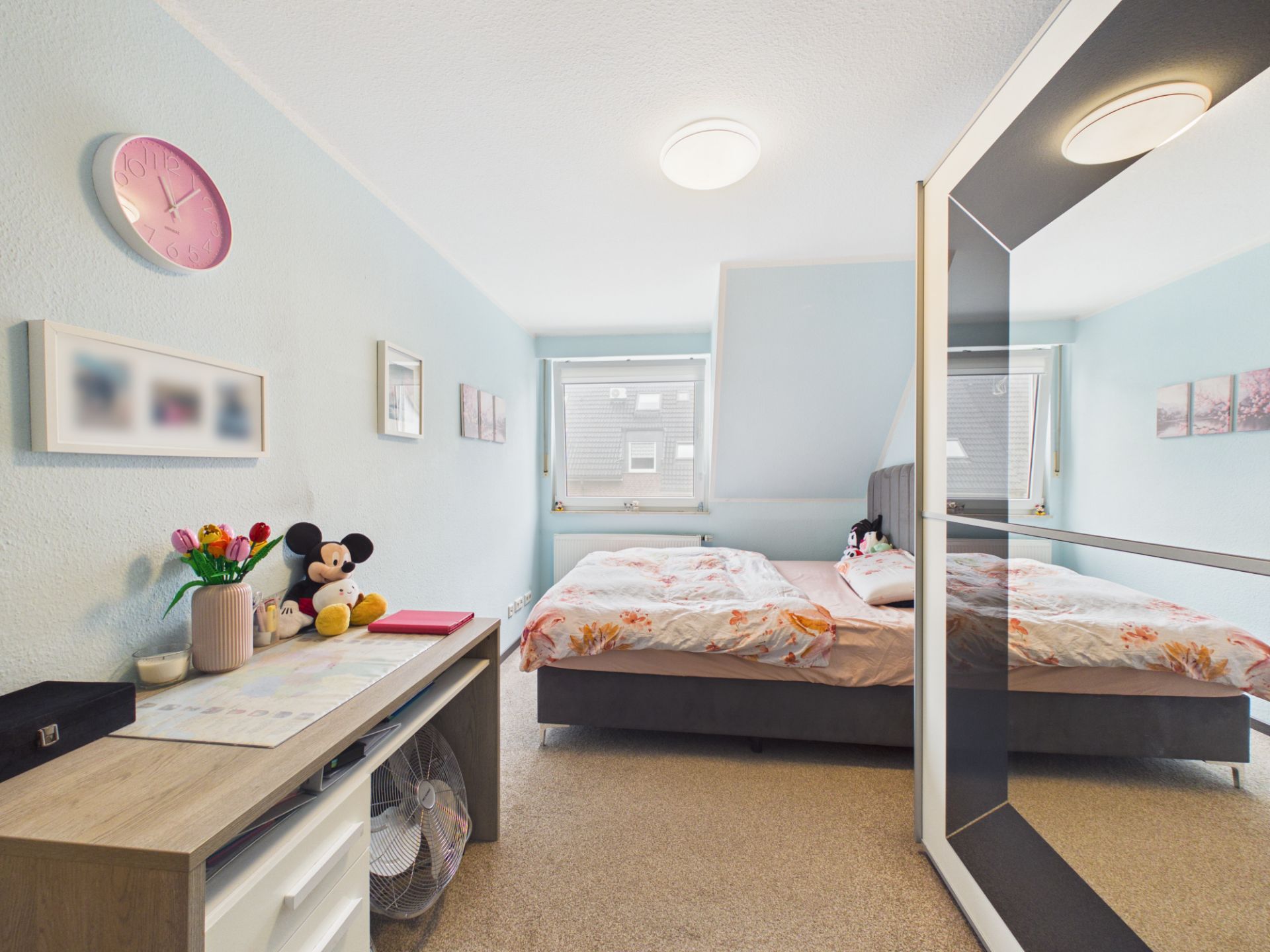 Helles Schlafzimmer mit Doppelbett, Schreibtisch, Spiegel, Dekoration und zwei Fenstern.