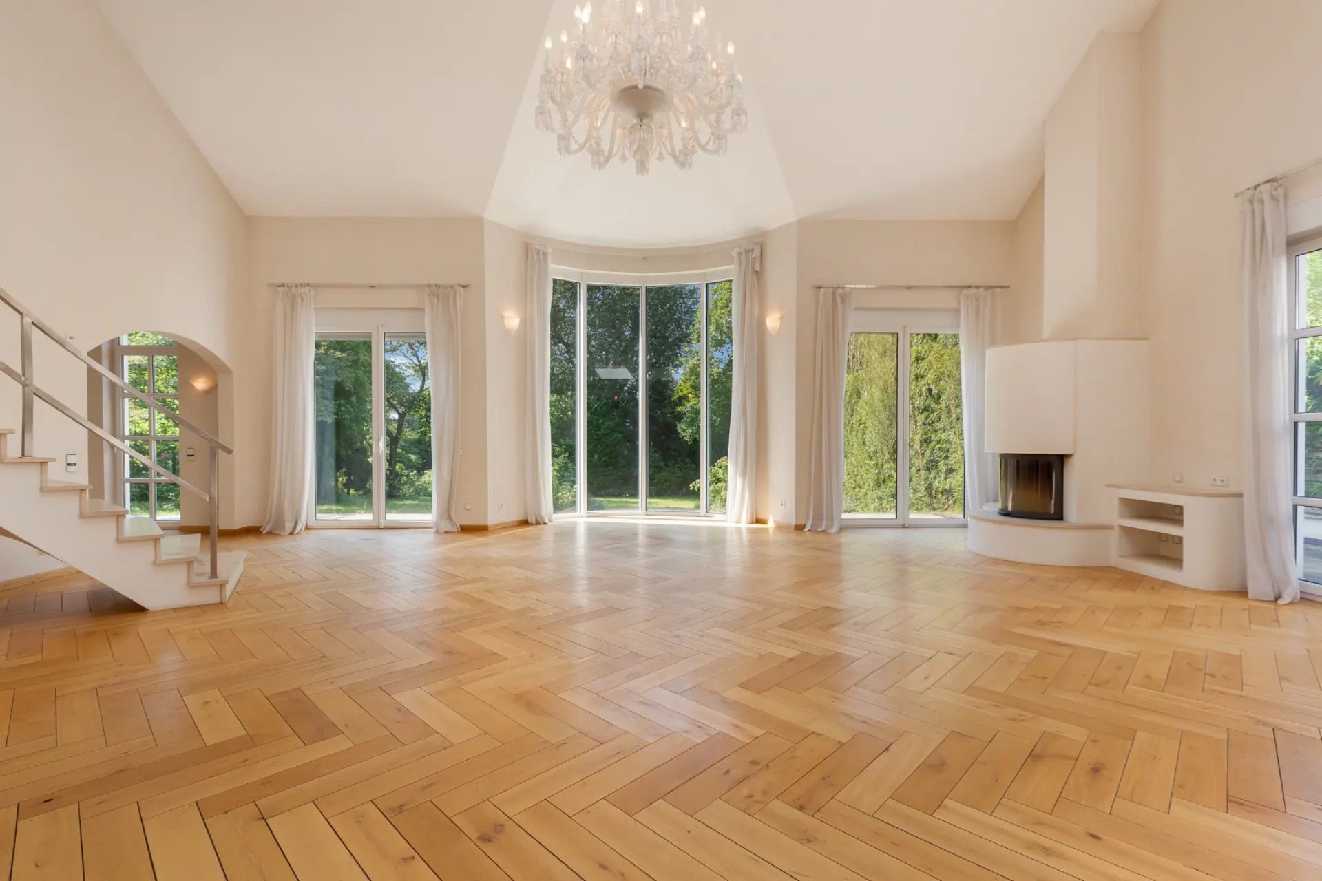 Helles Wohnzimmer mit Parkettboden, großen Fenstern, Kamin, Treppe und Kronleuchter.