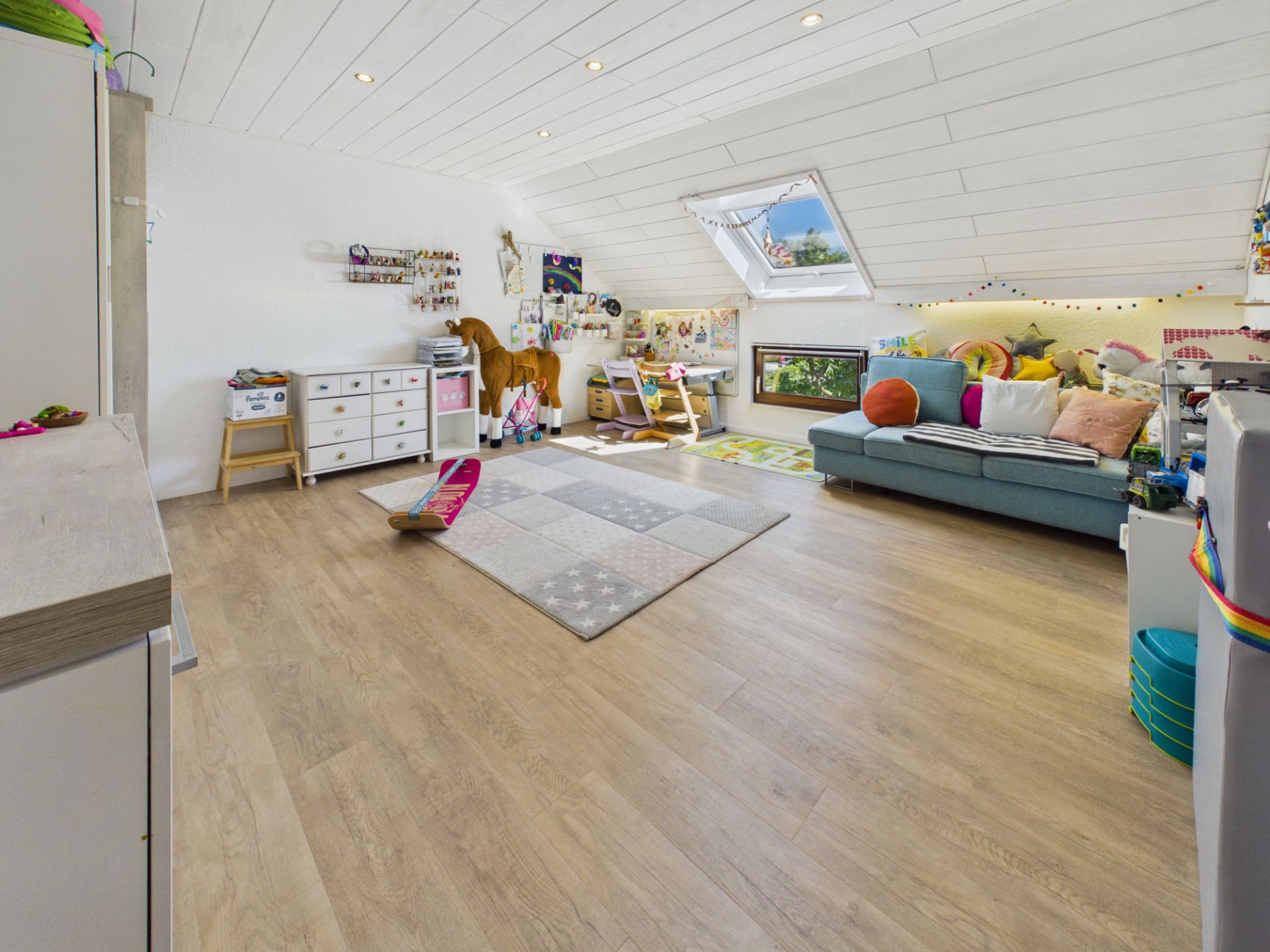 Helles Kinderzimmer mit Dachschrägen, Sofa, Teppich und Spielzeug auf Holzboden.