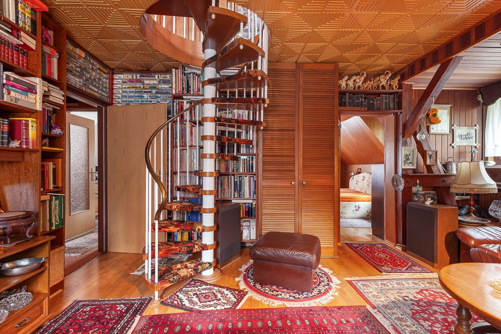 Wohnzimmer mit Holztreppe, Bücherregalen, Teppichen und einem braunen Hocker.