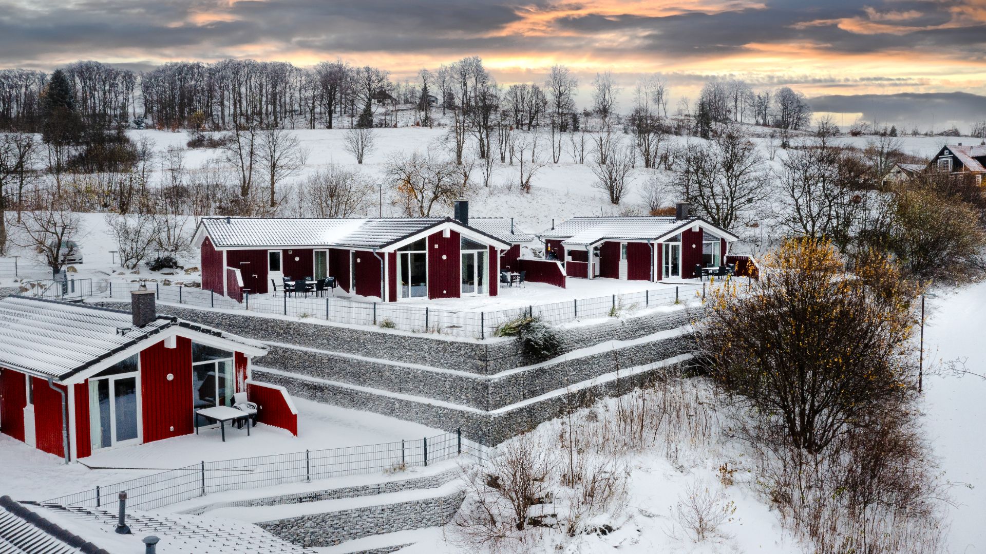 Rote Häuser mit weißen Dächern im verschneiten Garten, umgeben von Winterlandschaft und Bäumen.
