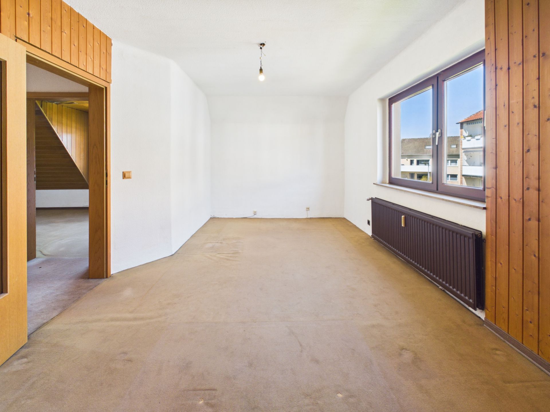 Leeres Zimmer mit braunem Teppich, großen Fenstern und Holzwandverkleidung.