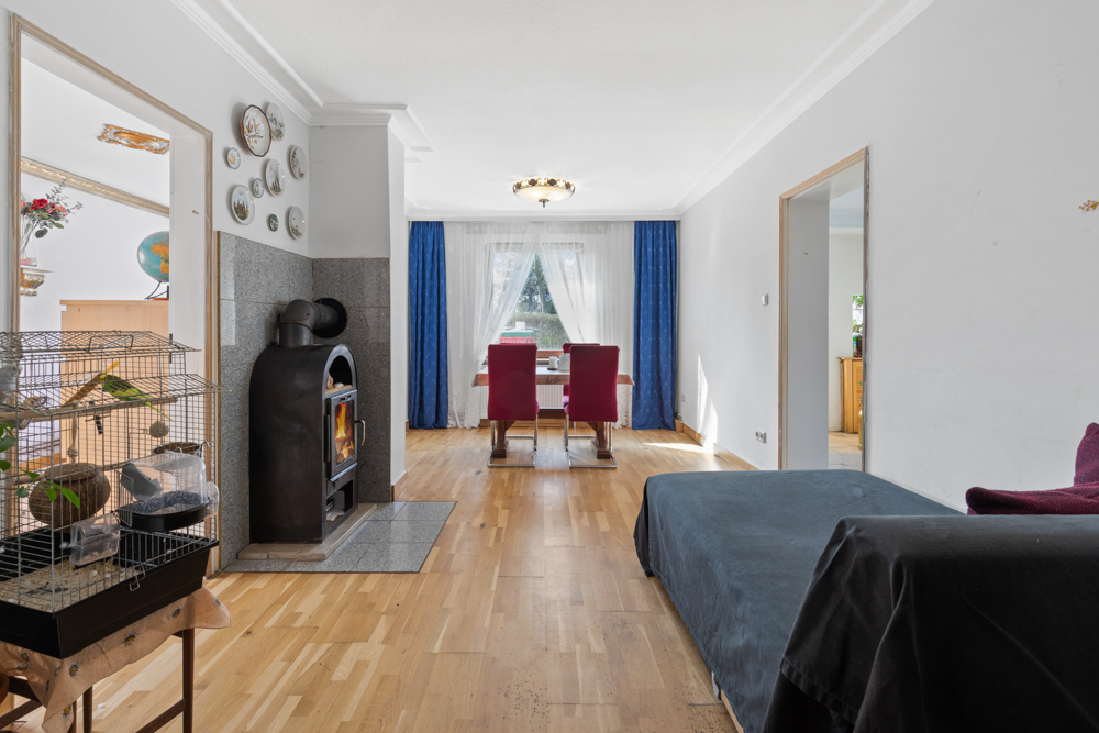 Wohnzimmer mit Holzofen, Vogelkäfig, Esstisch mit roten Stühlen und großen Fenstern mit blauen Vorhängen.