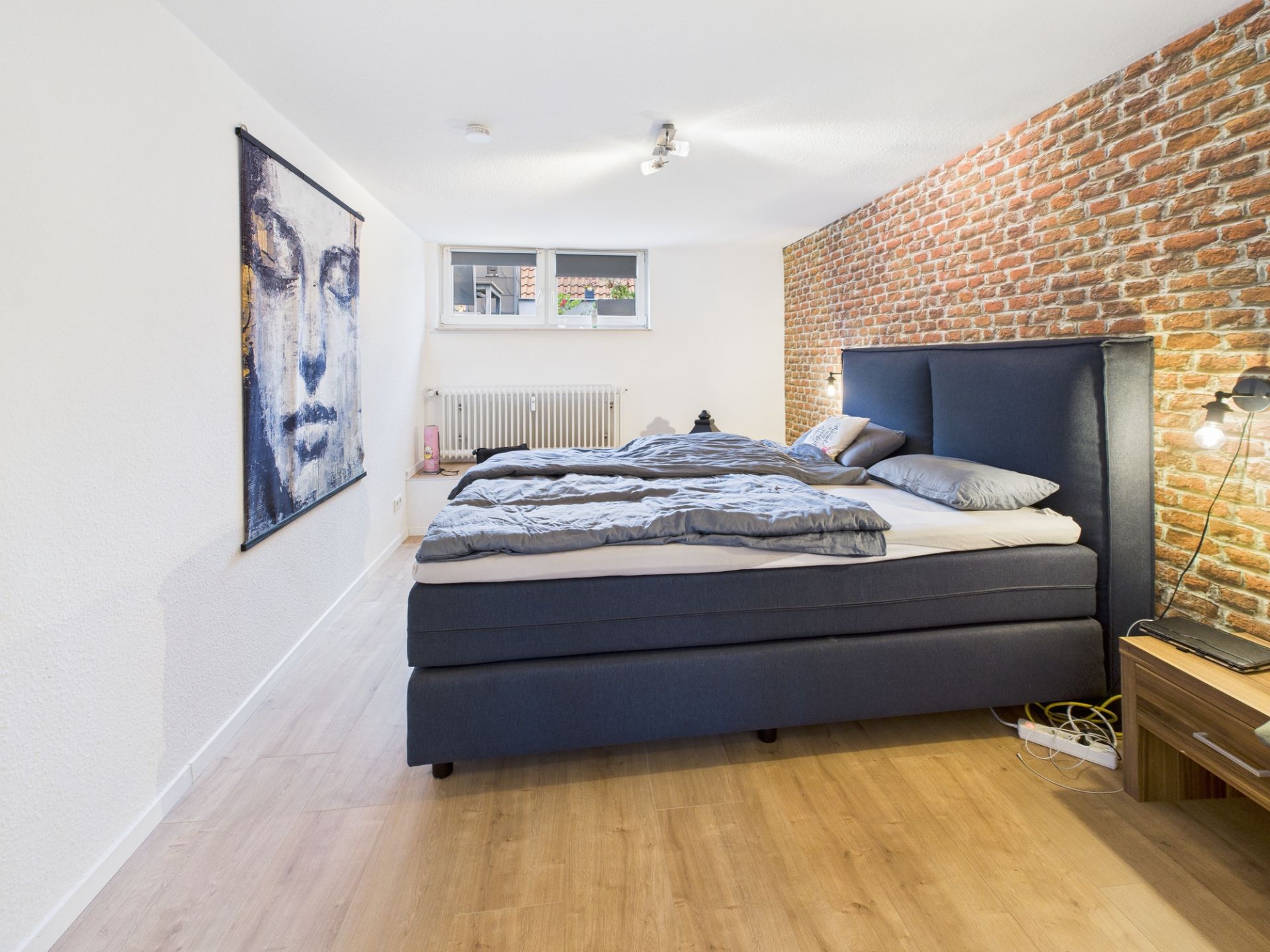 Schlafzimmer mit Doppelbett, Backsteinwand, Kunstbild und Fenster mit Heizung darunter.