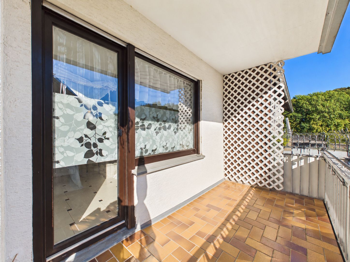 Balkon mit braunen Fliesen, großen Fenstern und weißem Sichtschutzgitter.