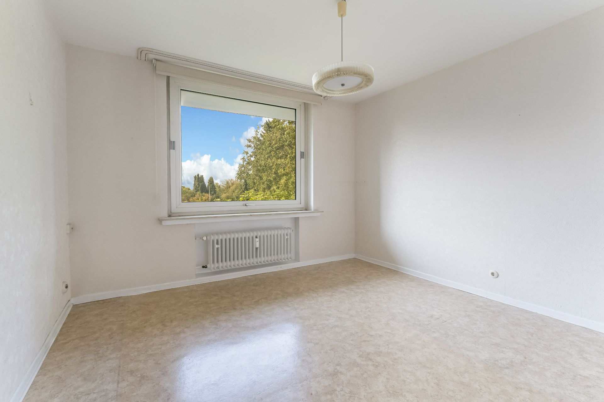 Leeres Zimmer mit großem Fenster, Heizung darunter und heller Deckenlampe.