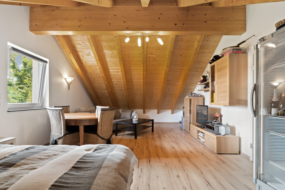 Gemütliches Dachzimmer mit Holzbalkendecke, Esstisch, Bett und Fernseher.