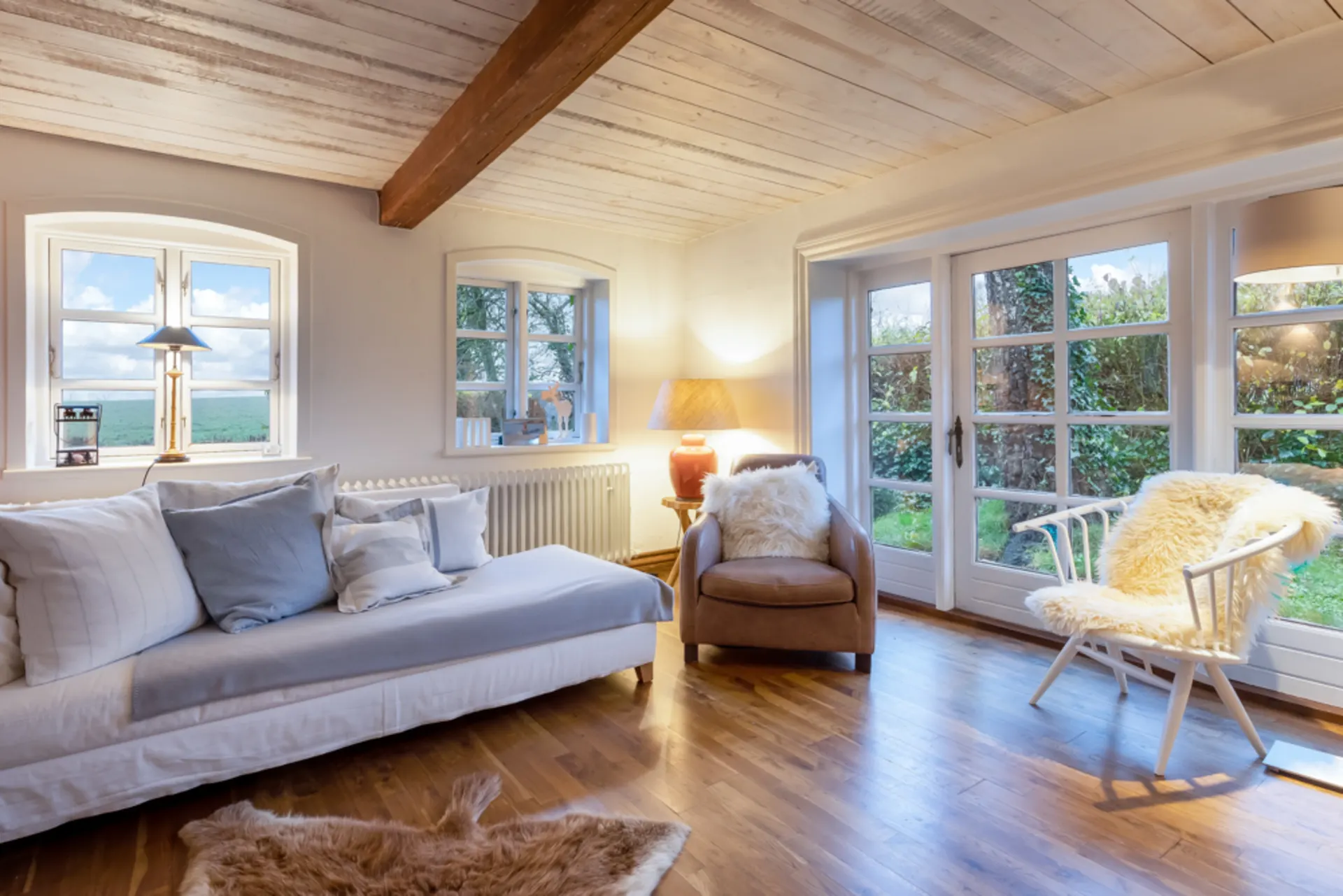 Helles Wohnzimmer mit Sofa, Sessel, Stuhl und großen Fenstern zum Garten.