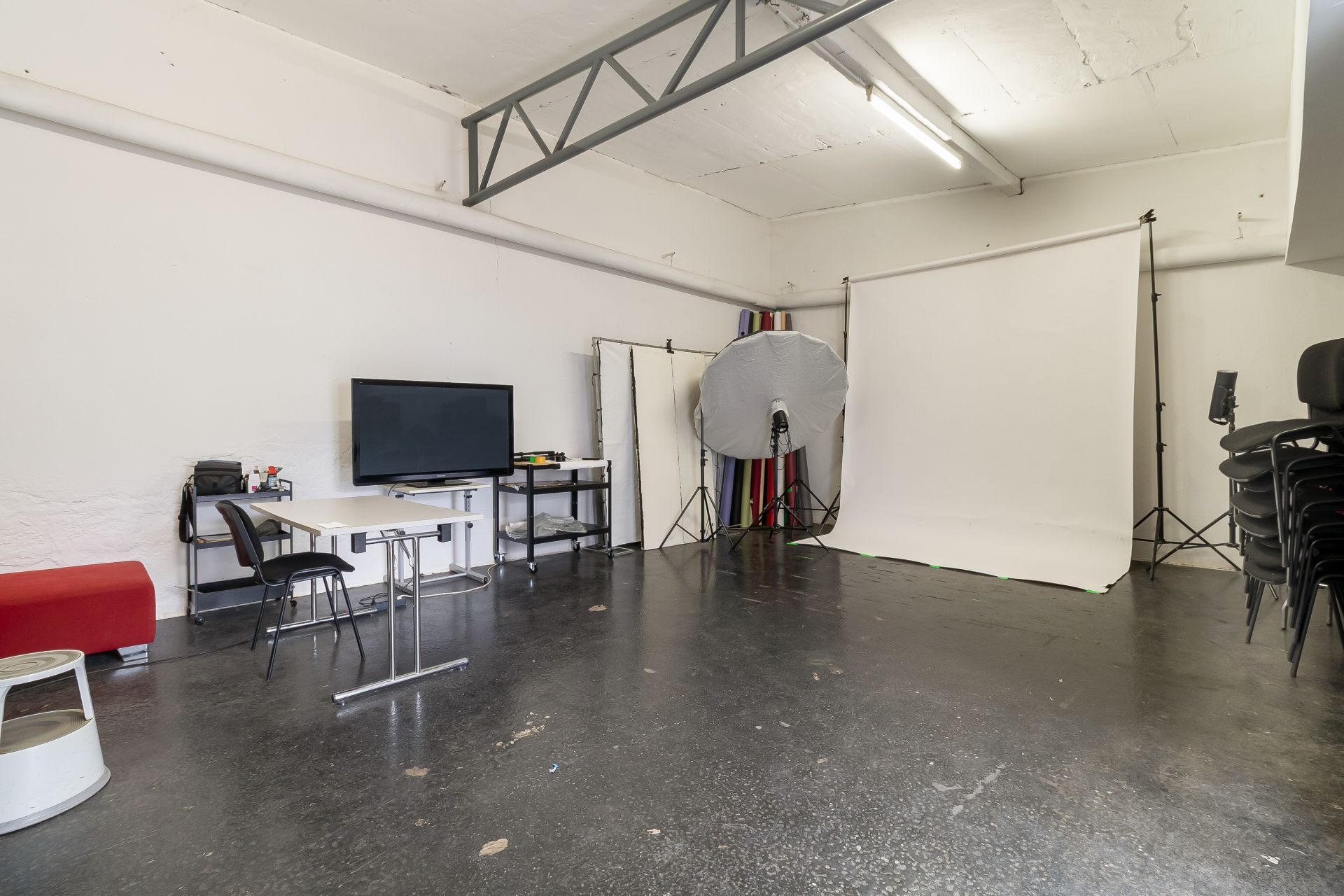 Fotostudio mit weißen Wänden, Fotografierhintergrund, Monitor, Tischen und Stühlen.