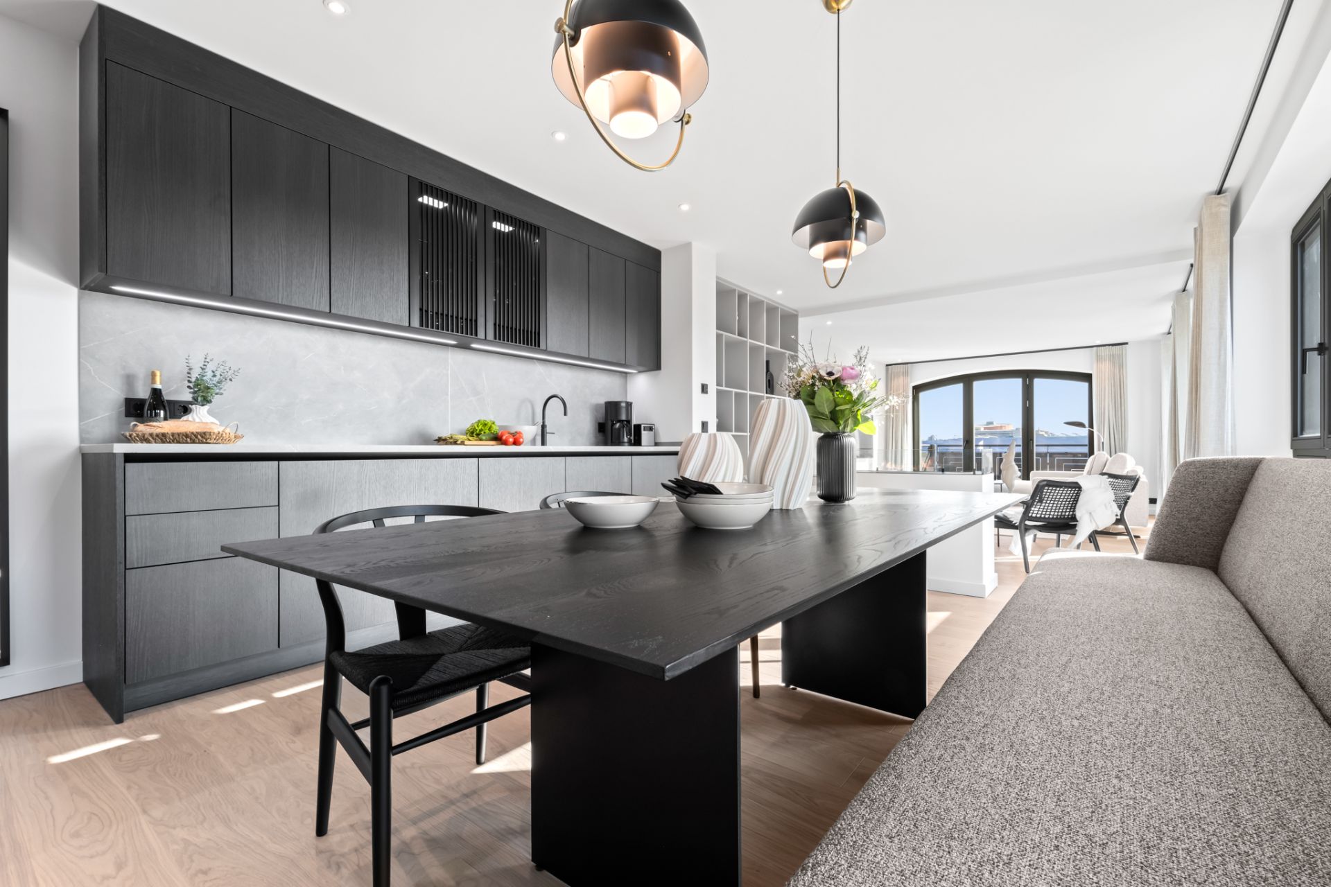 Moderne Küche mit dunklem Esstisch, Sofa, Stühlen und großen Fenstern in offenem Wohnraum.