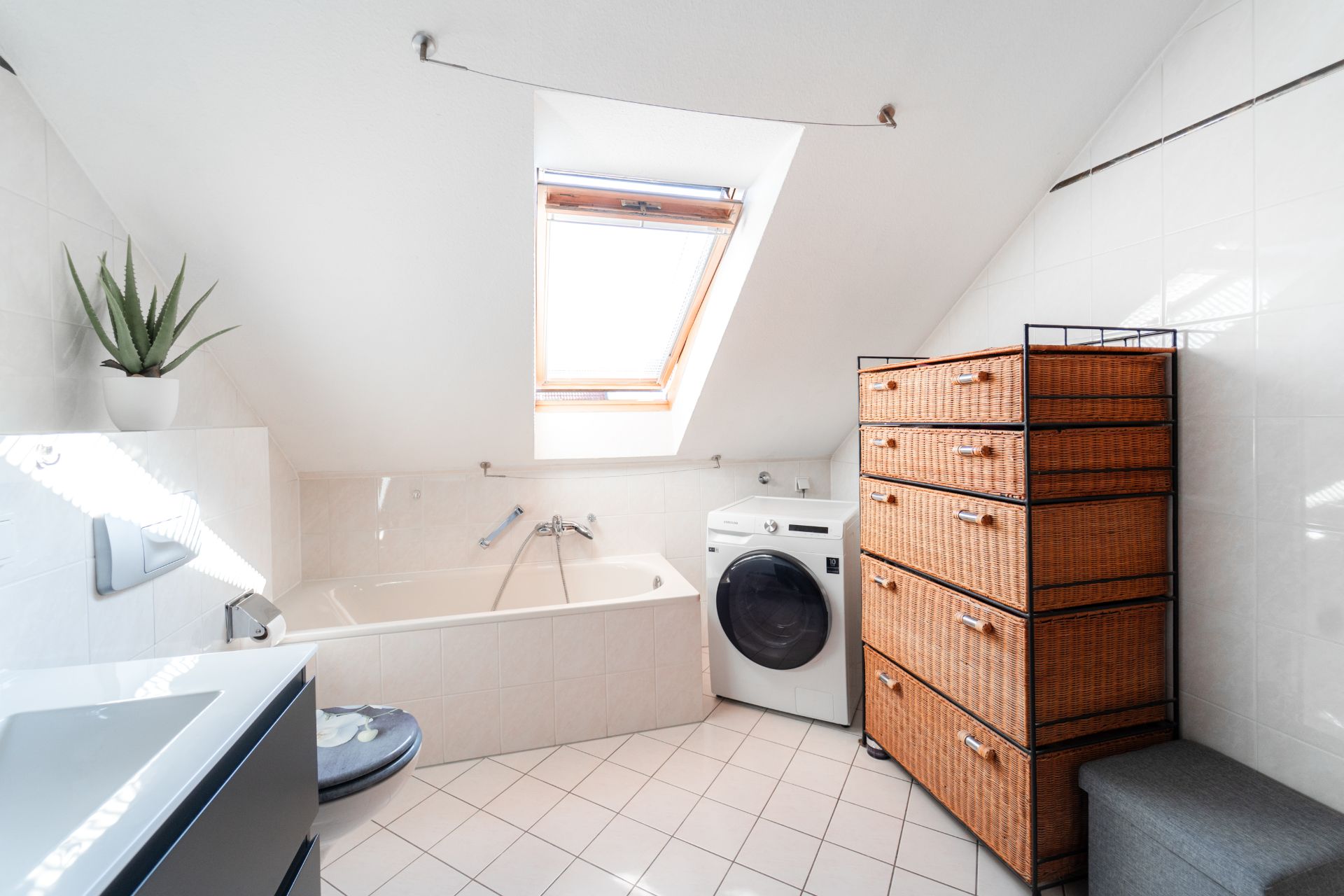 Helles Badezimmer mit Dachfenster, Badewanne, Waschmaschine und Schrank mit Körben.