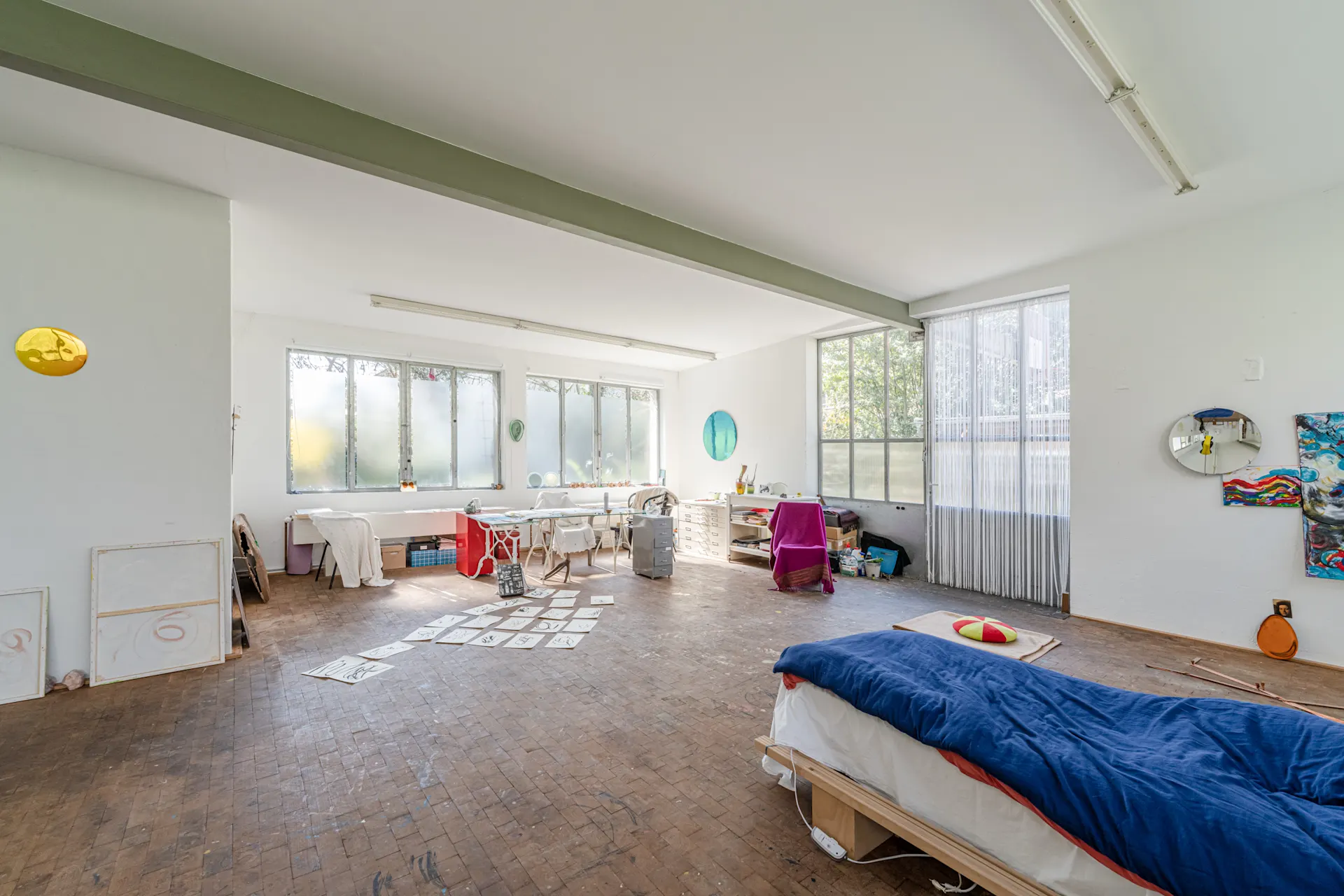 Heller Raum mit großem Fenster, Bett, Schreibtisch und Kunstmaterialien auf dem Boden.