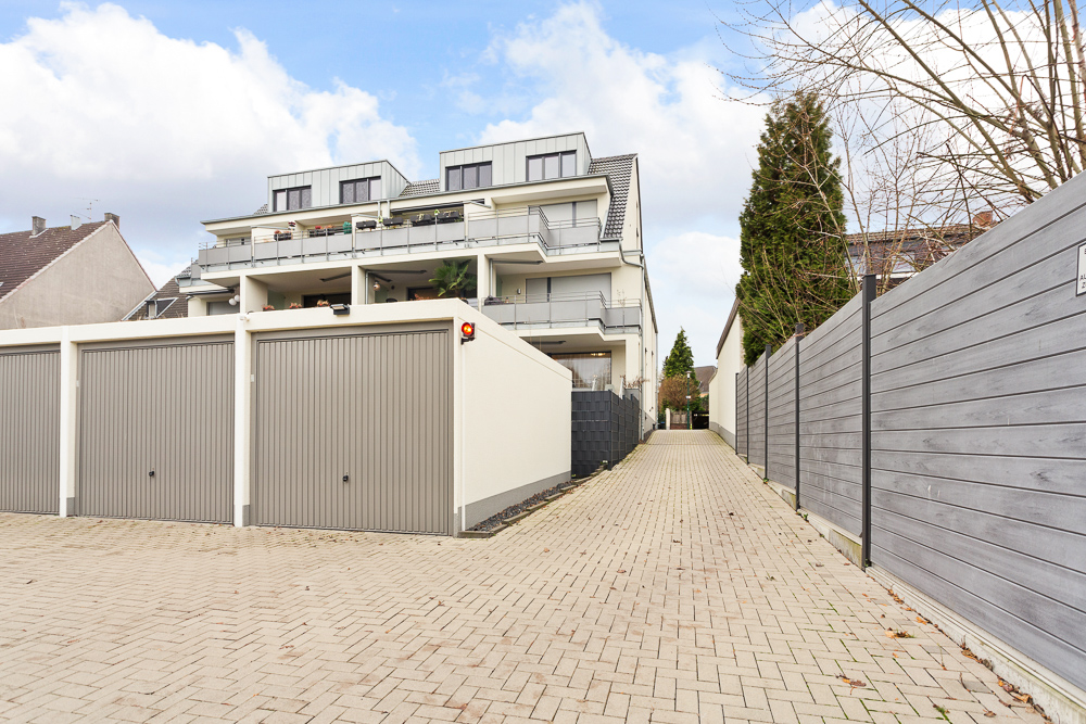 Mehrfamilienhaus mit mehreren Garagen und gepflasterter Einfahrt, daneben ein Holzzaun und Bäume.