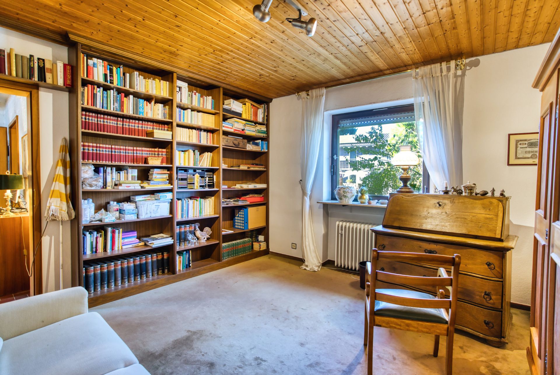 Helles Zimmer mit Bücherregal, Schreibtisch, Stuhl und Fenster mit Vorhängen.