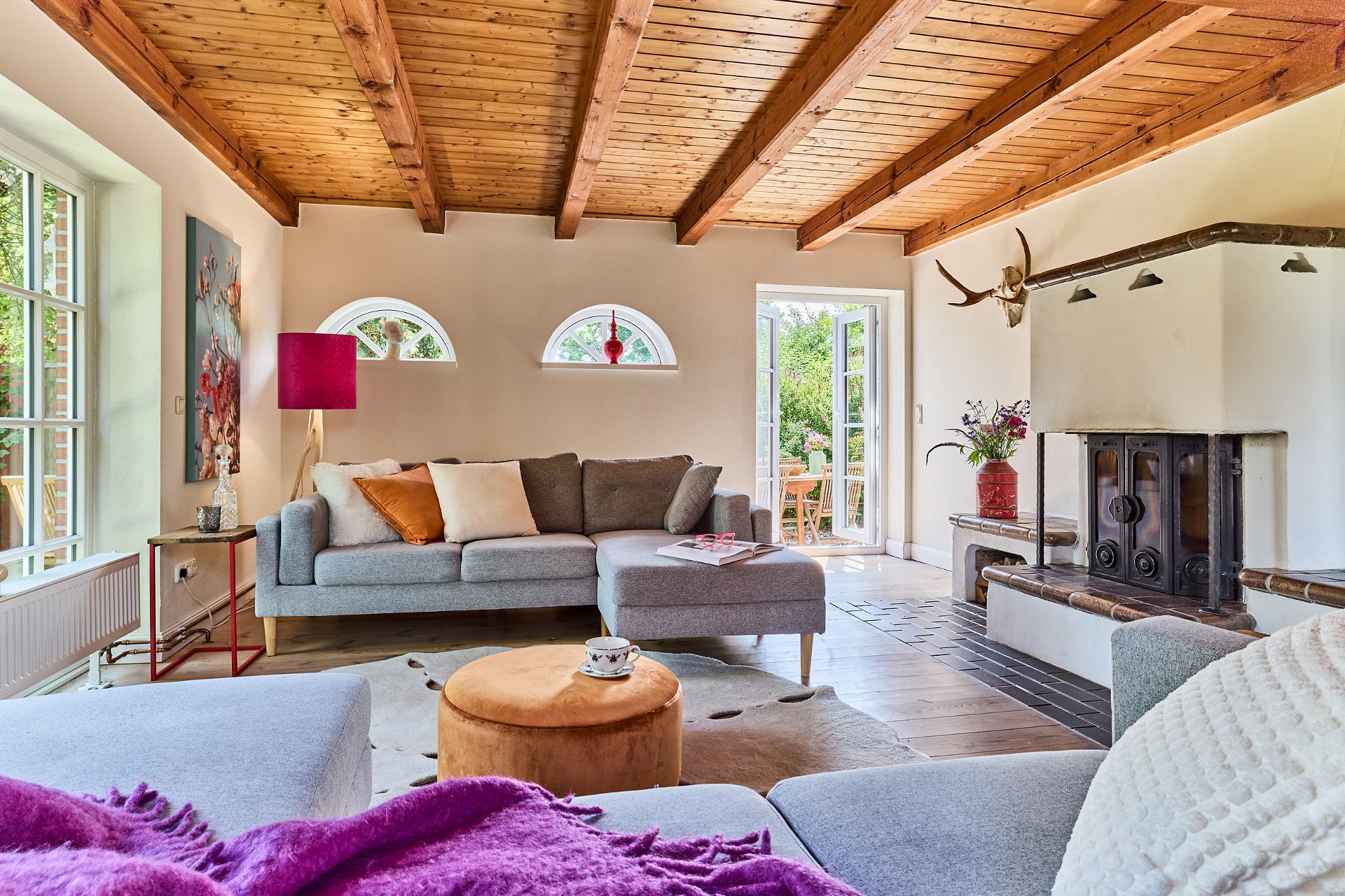 Helles Wohnzimmer mit Holzbalkendecke, Ecksofa, Kaminofen und Zugang zur Terrasse.