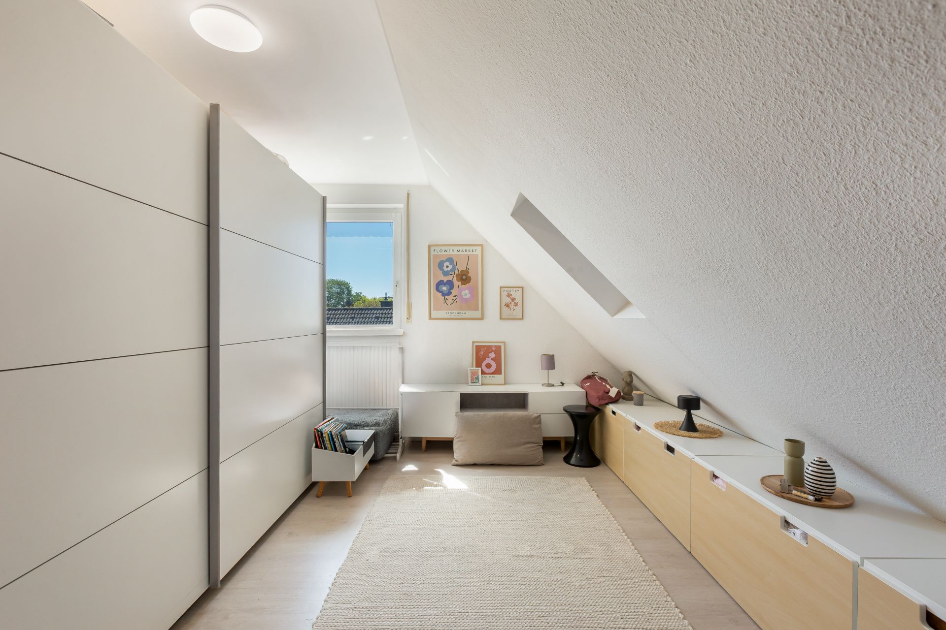 Helles Zimmer mit Schräge, kleinem Sofa, Schränken, Teppich und Wandbildern.
