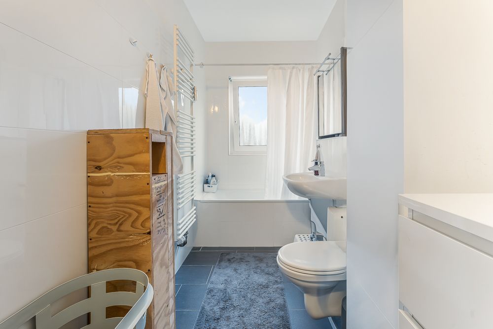 Helles Badezimmer mit Badewanne, Waschbecken, Toilette, Holzregal und Fenster mit Vorhang.